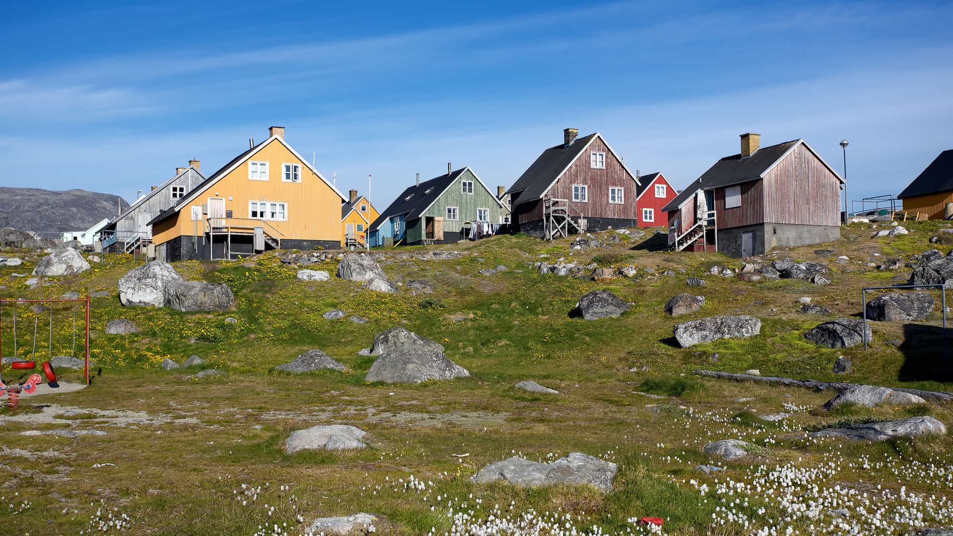A row of brightly colored wooden houses in the village of Nanortalik, Greenland. The houses, painted in red, blue, green, and yellow, are built on a rocky, grassy slope. There are large boulders scattered across the landscape, and patches of white flowers dot the ground. In the background, rolling hills rise under a clear blue sky with thin clouds. The scene captures the unique architecture and rugged landscape of Greenland, an expedition destination.
