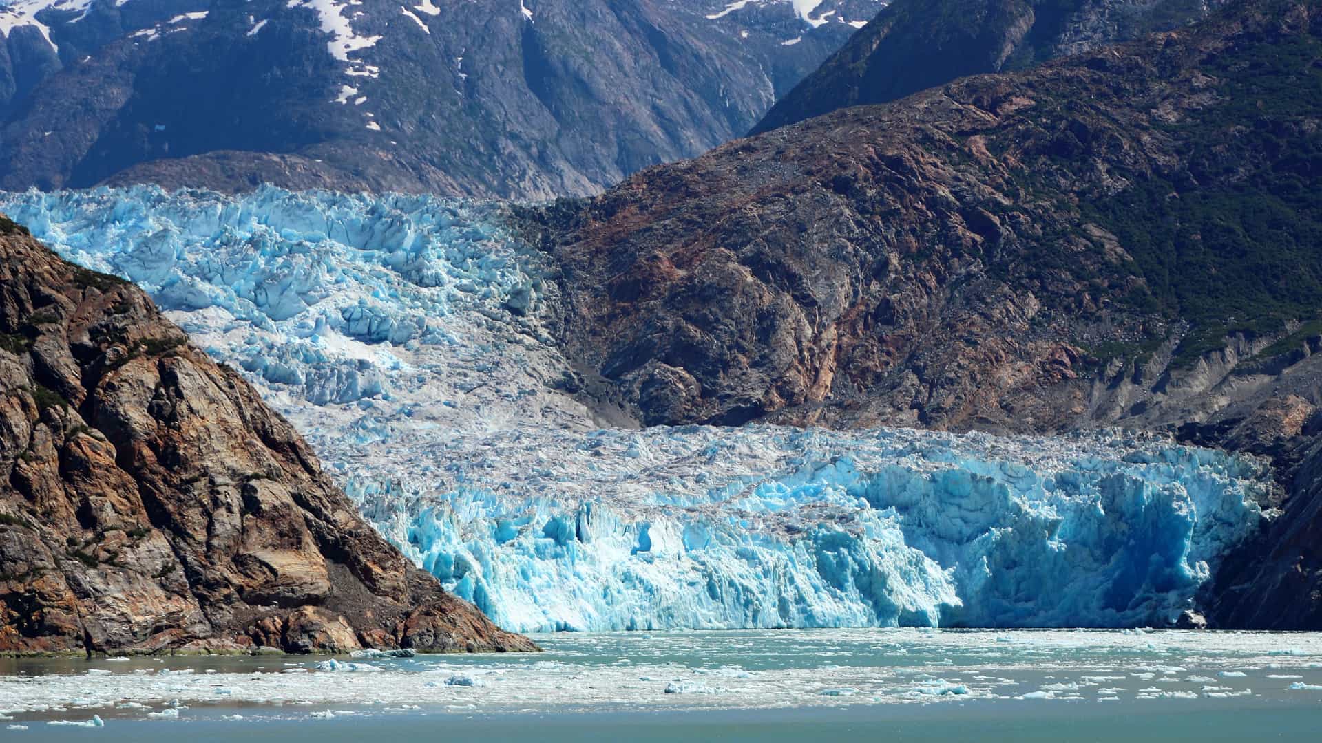 A striking view of the Sawyer Glacier in the Tracy Arm Fjord, Alaska. The glacier is a massive wall of brilliant blue and white ice, with deep crevasses and a rough, textured surface. It is nestled between towering, rocky mountains, some of which are still covered in snow. The base of the glacier meets a body of water filled with floating ice chunks. The image captures the immense scale and raw beauty of this icy landscape in the Alaska region.
