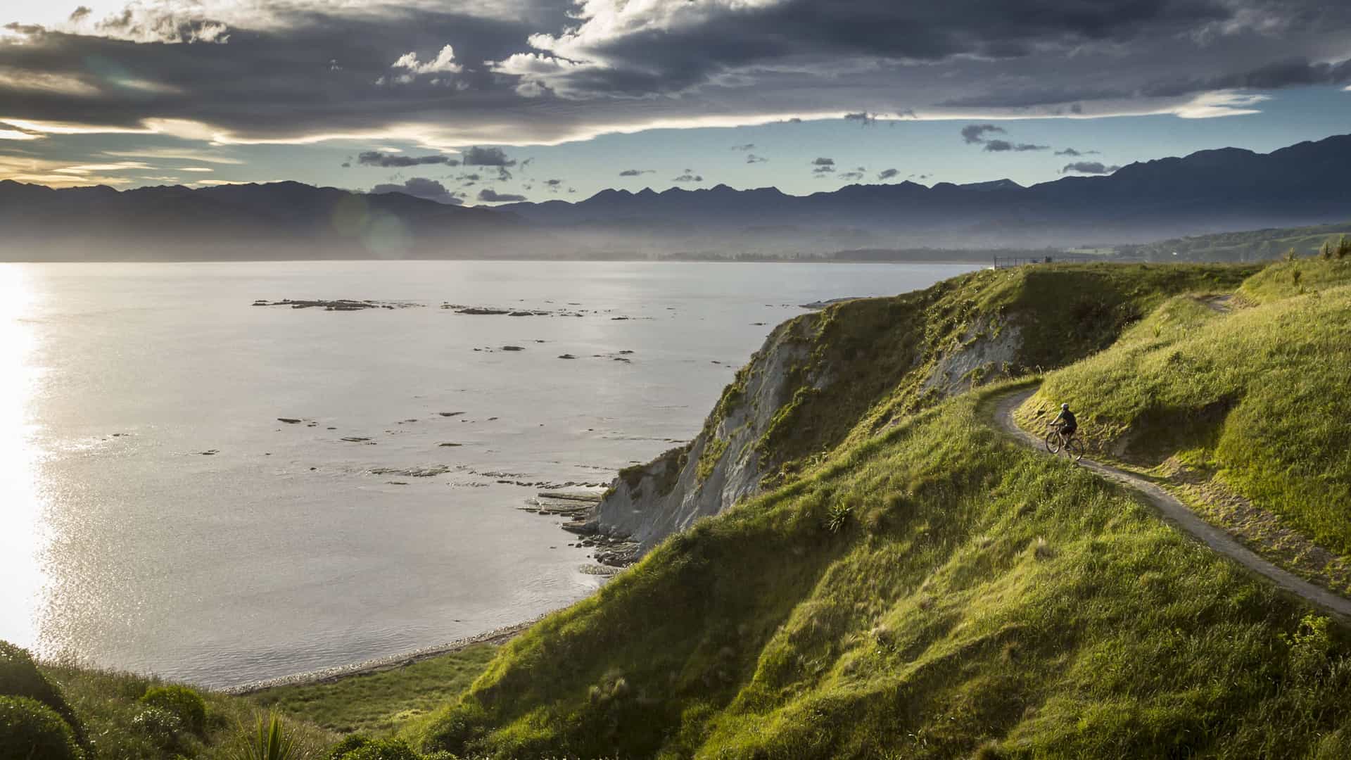 A beautiful sunset view of the coastline near Kaikoura, New Zealand. A winding path cuts through the bright green, grassy hill in the foreground. The hill descends to a rocky shoreline and a calm body of water, where the setting sun creates a brilliant, shimmering reflection. In the distance, a line of dark mountains fades into the horizon under a dramatic, cloudy sky. The image captures the diverse and stunning landscape of New Zealand's coastline.