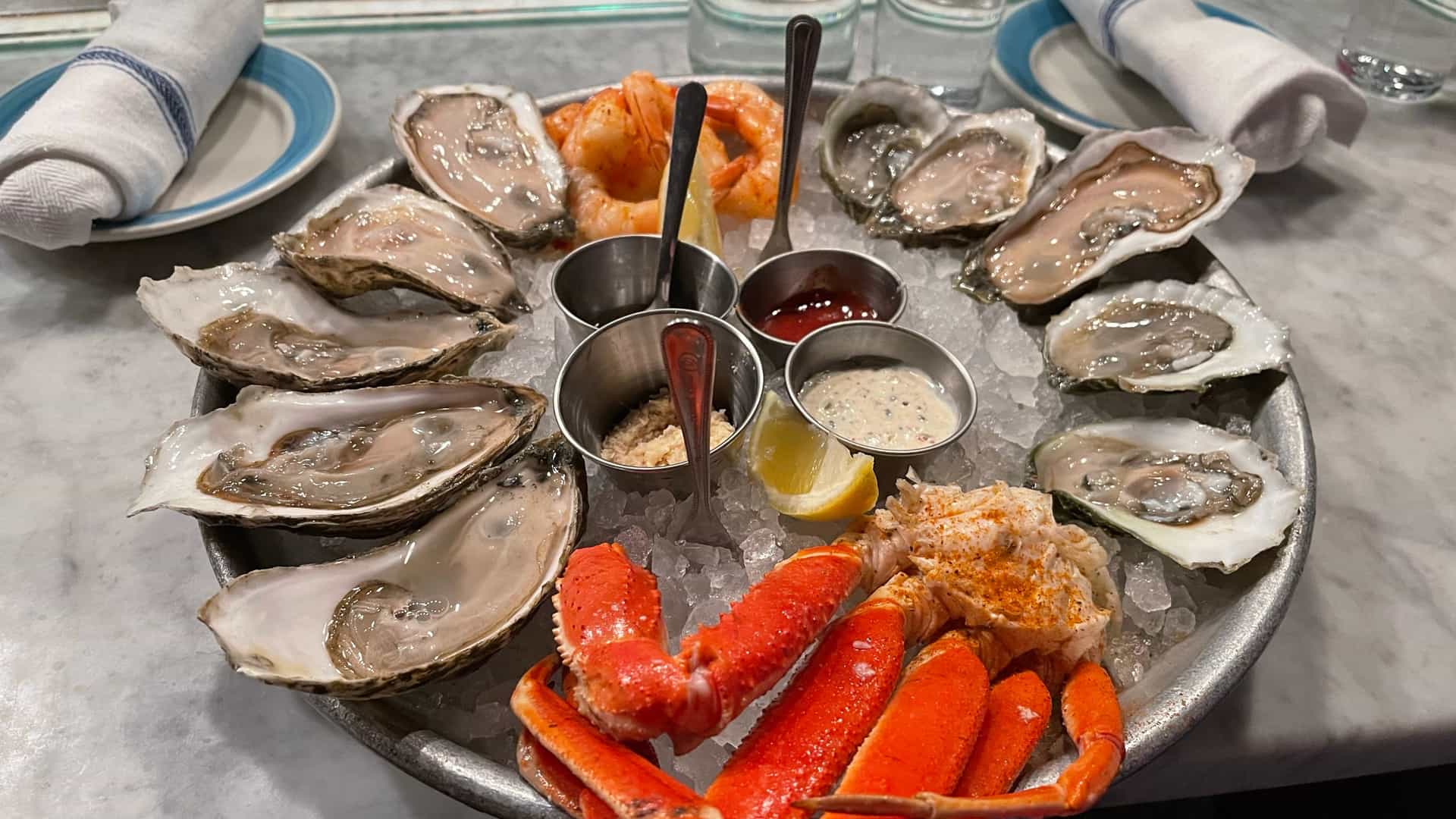 A large metal platter filled with fresh seafood on ice, including raw oysters, snow crab legs, and shrimp, served with dipping sauces and lemon on a marble tabletop.