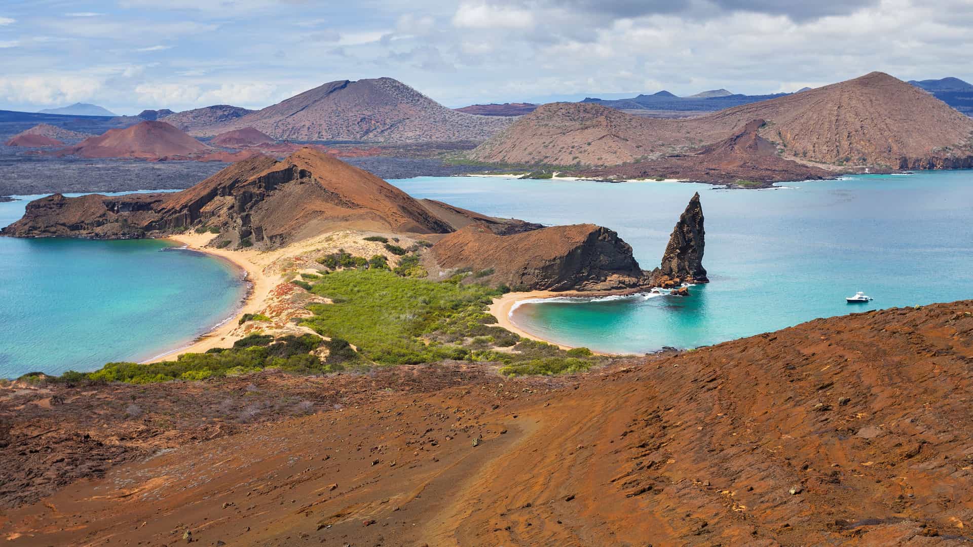An iconic view of Bartolomé Island in the Galapagos Islands, Ecuador. The image shows the famous Pinnacle Rock, a volcanic rock formation, rising sharply from the turquoise-blue water of a crescent-shaped bay. The surrounding landscape is a mix of volcanic terrain with reddish-brown soil and sparse green vegetation. In the background, other volcanic cones are visible, showcasing the unique geological features of the Galapagos Islands region.