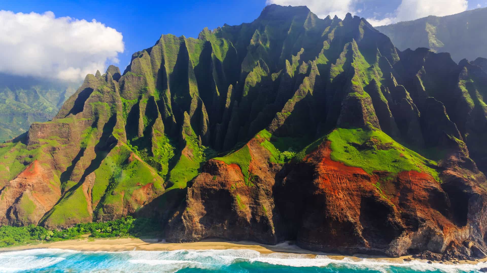 Dramatic, deeply ridged green and reddish-brown mountain cliffs rise steeply above a sandy beach and turquoise ocean waves along the coast.