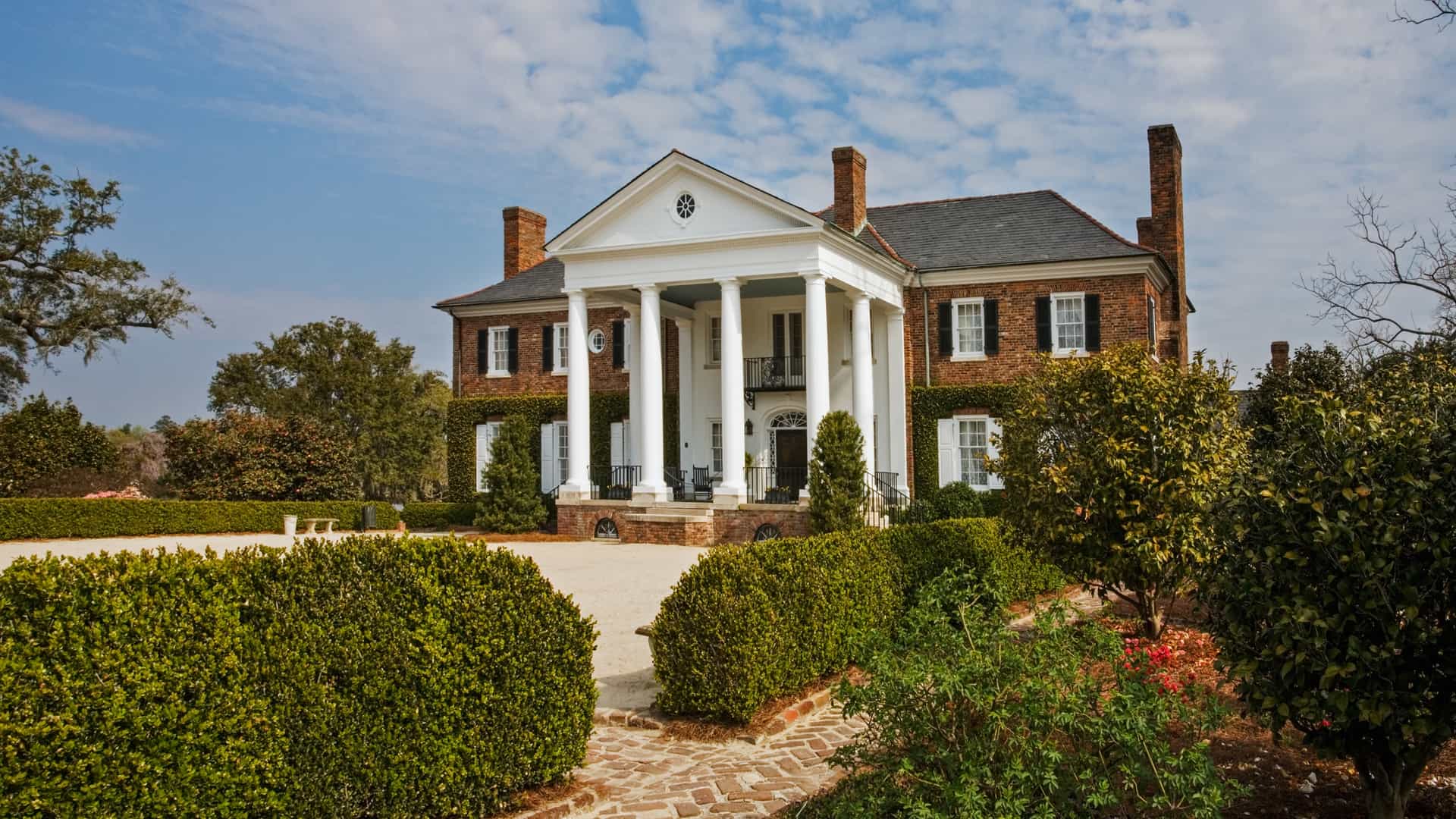 A grand two-story brick mansion featuring iconic white Neoclassical columns, manicured green hedges, and a historic brick-paved walkway under a clear blue sky.