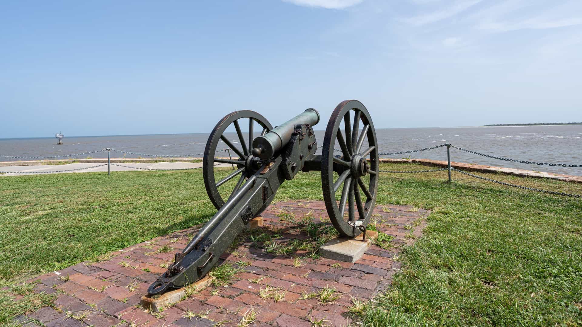 A historic green-tinted cannon mounted on a carriage sits on a brick platform in a grassy coastal park overlooking the open water.