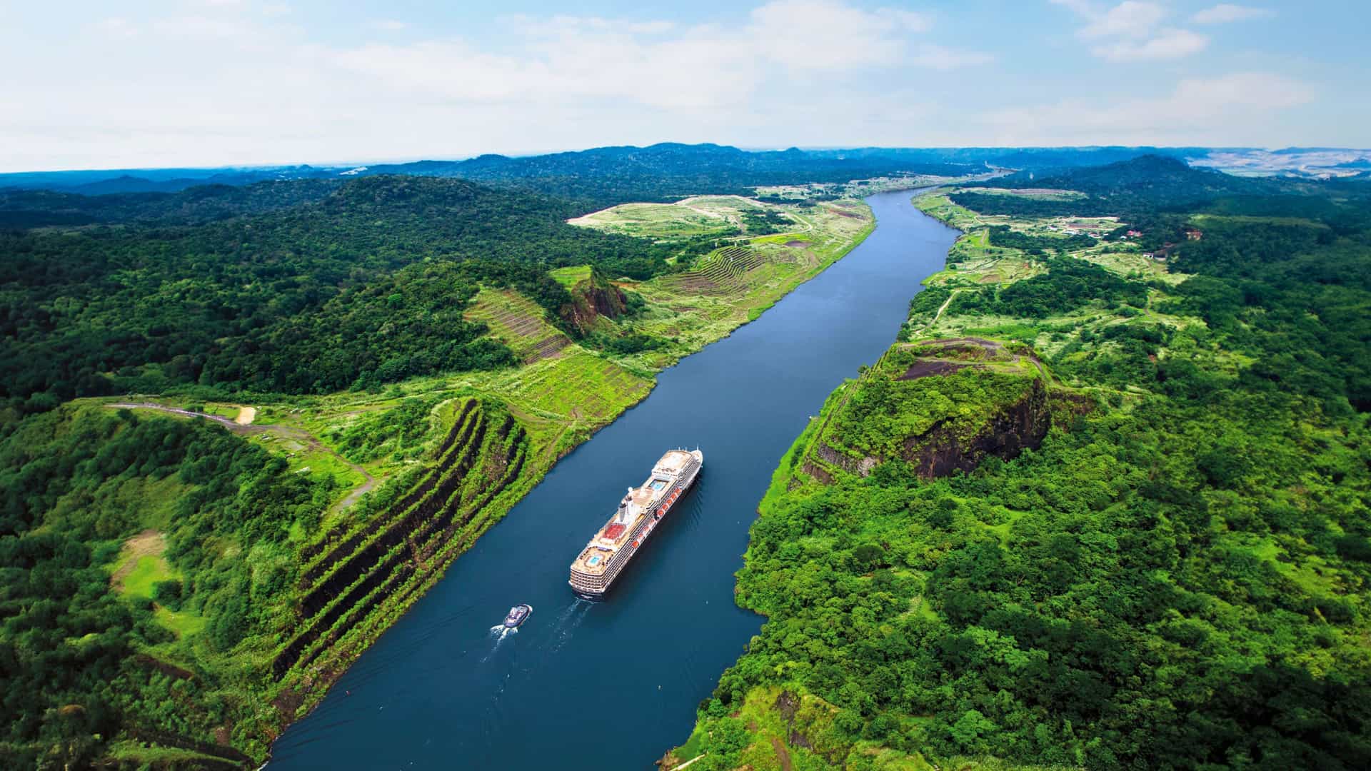 Holland America Cruise Experience: Aerial view of their majestic ship transiting the iconic Panama Canal, framed by lush rainforest and mountains under blue sky.