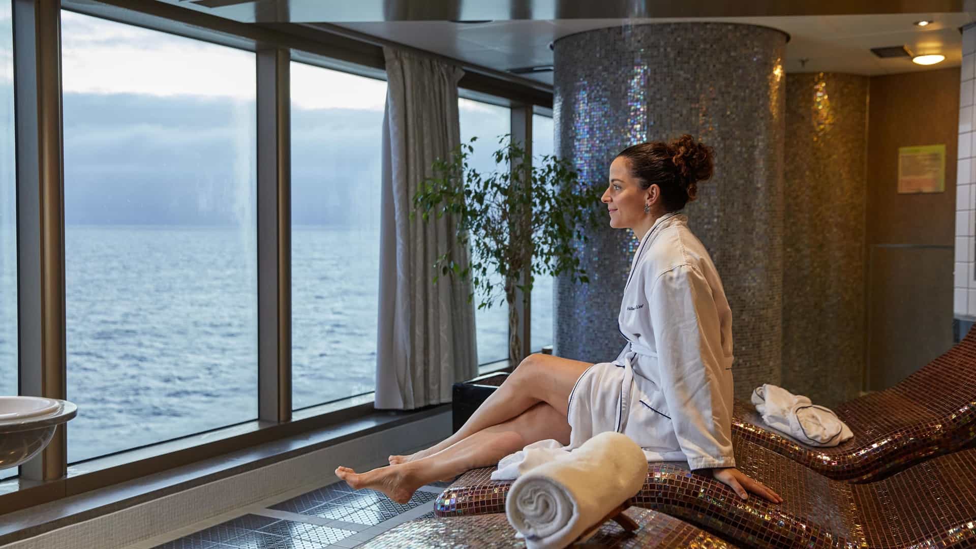 A woman on the Holland America Line's Volendam ship is sitting on a heated ceramic lounger in the Greenhouse Spa, wearing a white bathrobe and looking out at the ocean through large windows.