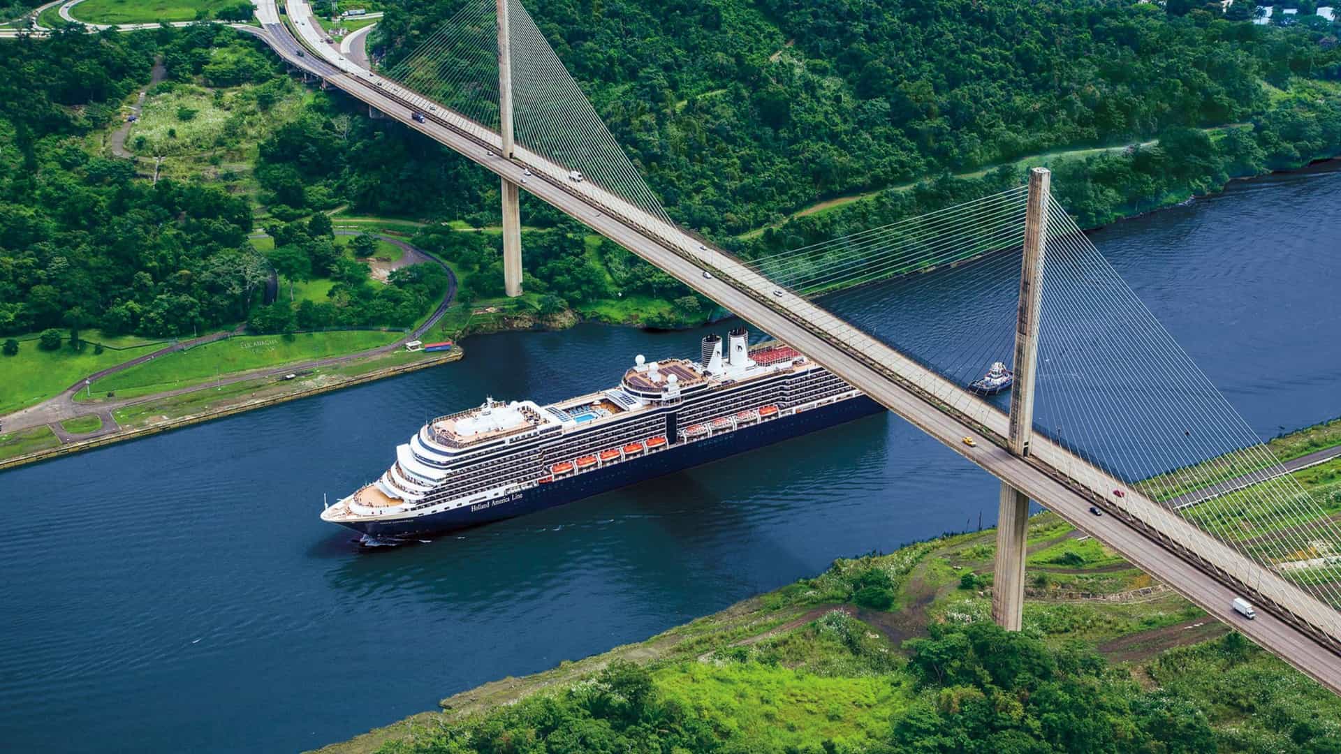 A large Holland America Line cruise ship sails under a massive cable-stayed bridge through the narrow waters of the Panama Canal, surrounded by lush green tropical vegetation.
