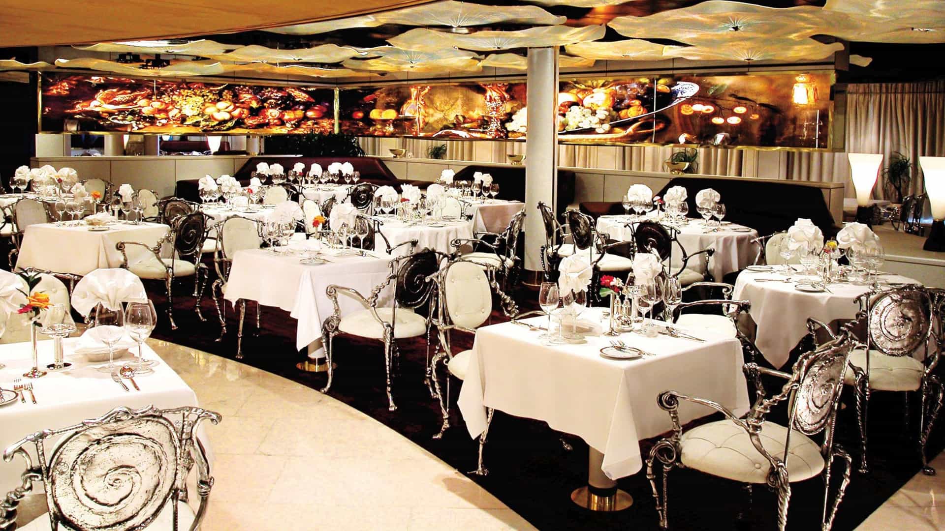 An elegant fine dining room featuring tables set with white tablecloths, glassware, and ornate silver chairs, beneath warm ambient lighting and a illuminated ceiling feature.