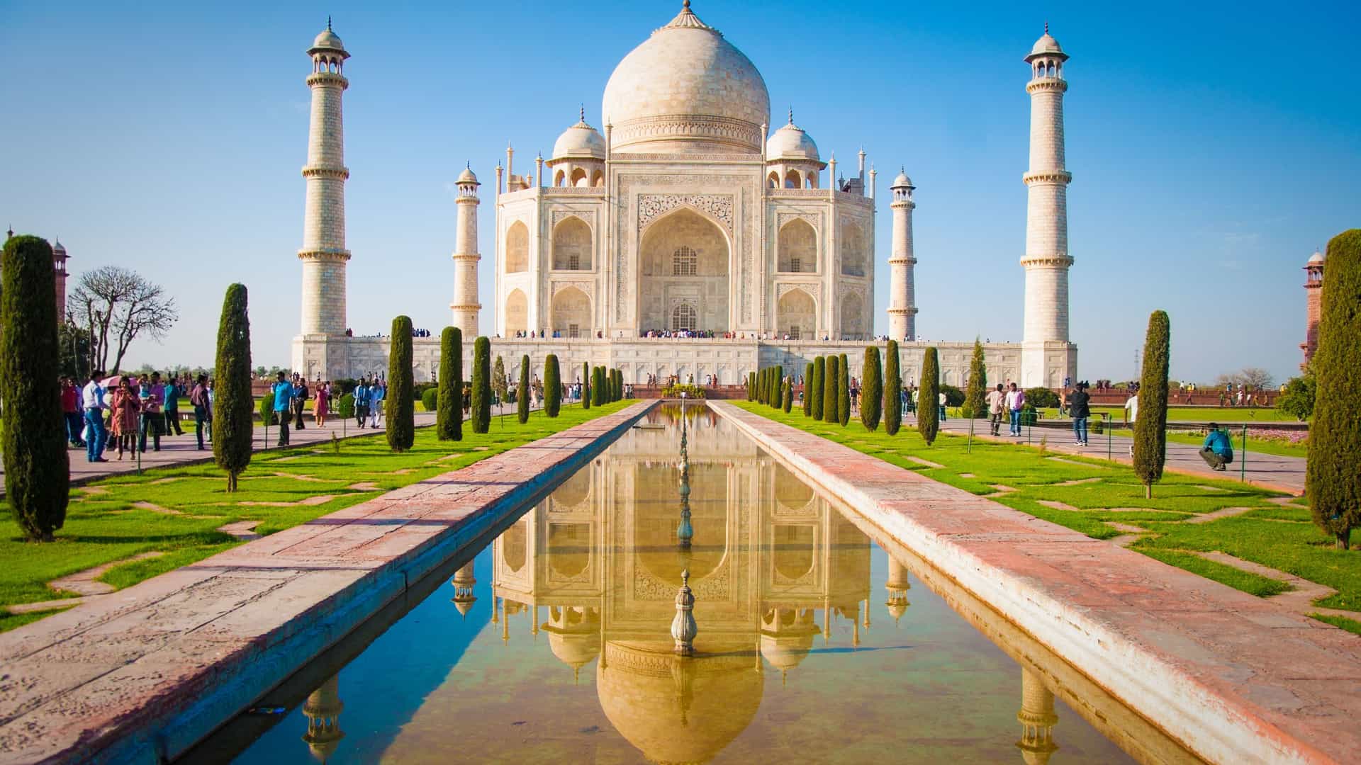 The majestic Taj Mahal in Agra, India, with its white marble mausoleum and surrounding minarets, is perfectly reflected in a long, rectangular pool of water. The symmetrical architecture is highlighted under a clear, brilliant blue sky. The foreground features a well-manicured green lawn with rows of tall, slender trees and a few visitors. The image captures the grandeur and beauty of this iconic landmark of the India region.