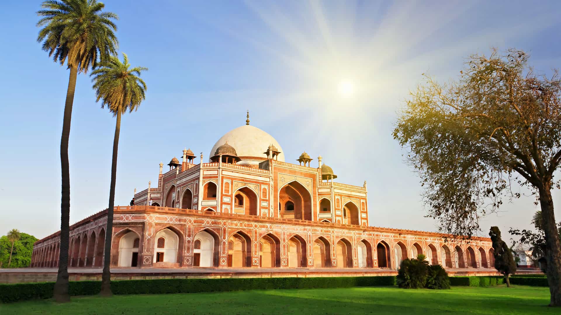The majestic Humayun's Tomb in New Delhi, India, bathed in the warm light of the setting sun. The building's red sandstone and white marble architecture is surrounded by a lush green lawn. The central dome and arches are prominent, and two tall palm trees are visible in the foreground. The sun, a bright starburst, is high in the sky, highlighting the beauty of this historical landmark of the India region.