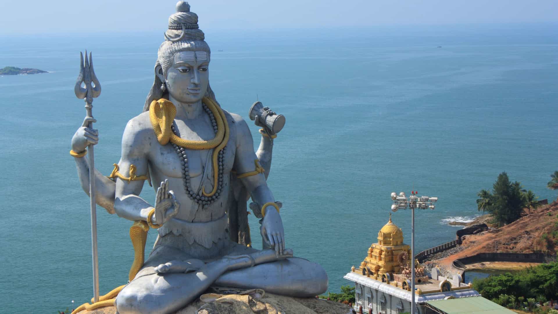 The towering statue of Lord Shiva in Murudeshwar, Karnataka, India, sits in a meditative pose overlooking the Arabian Sea. The large, silver-colored statue has a golden cobra around its neck and holds a trident in one hand and a drum in the other. In the foreground, a small, golden temple with a tiered roof is visible on the rocky coastline. The vast blue ocean stretches to the horizon under a bright, clear sky, capturing the spiritual and scenic beauty of the India region.