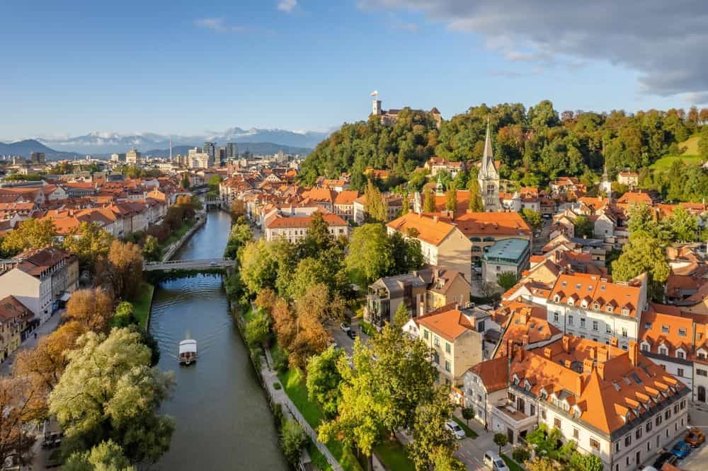 Aerial view of Ljubljana old town in autumn, featuring Ljubljana Castle, historic architecture, and the Ljubljanica River with the Slovenian Alps in the background on a sunny day