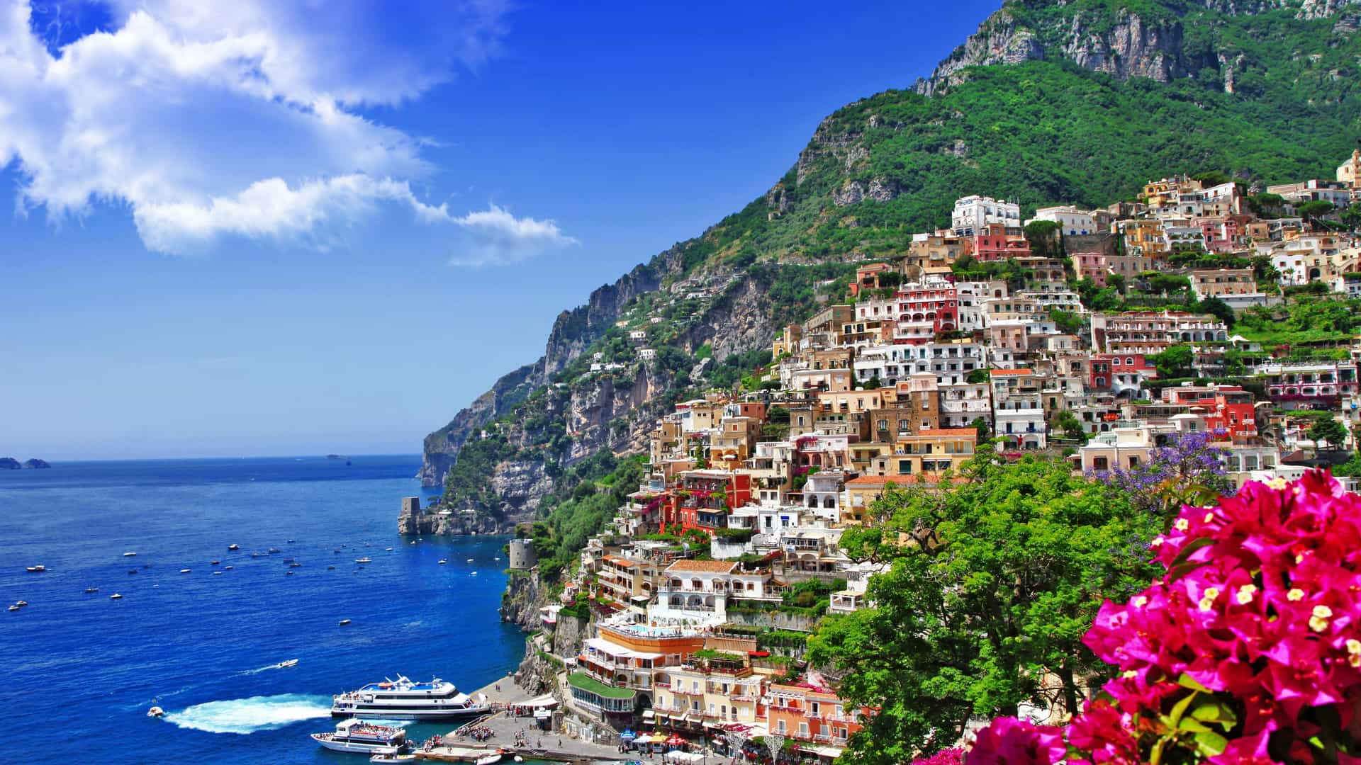 An iconic view of the seaside village of Positano on the Amalfi Coast in Italy. Colorful buildings are built into the steep cliffside, overlooking the turquoise Mediterranean Sea. Boats are scattered across the water, and a small beach is visible at the base of the cliffs. The lush green mountainside rises dramatically behind the village, and the sky is a brilliant blue with scattered white clouds. The image beautifully captures the unique and picturesque landscape of this Mediterranean region.