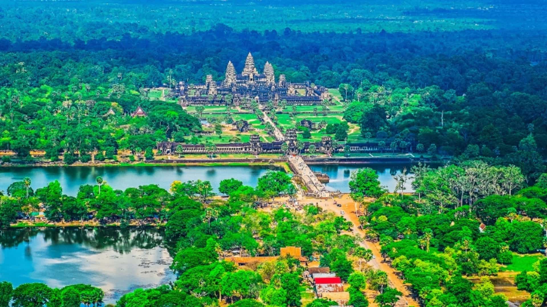 An aerial view of the majestic Angkor Wat temple complex in Cambodia, near the Mekong River. The ancient temple's towers and surrounding structures are visible within a vast expanse of dense, green forest. In the foreground, a large body of water, part of the temple's moat, reflects the sky, with a stone causeway leading across it. The image beautifully captures the scale and historical significance of this landmark within the lush Southeast Asian landscape.