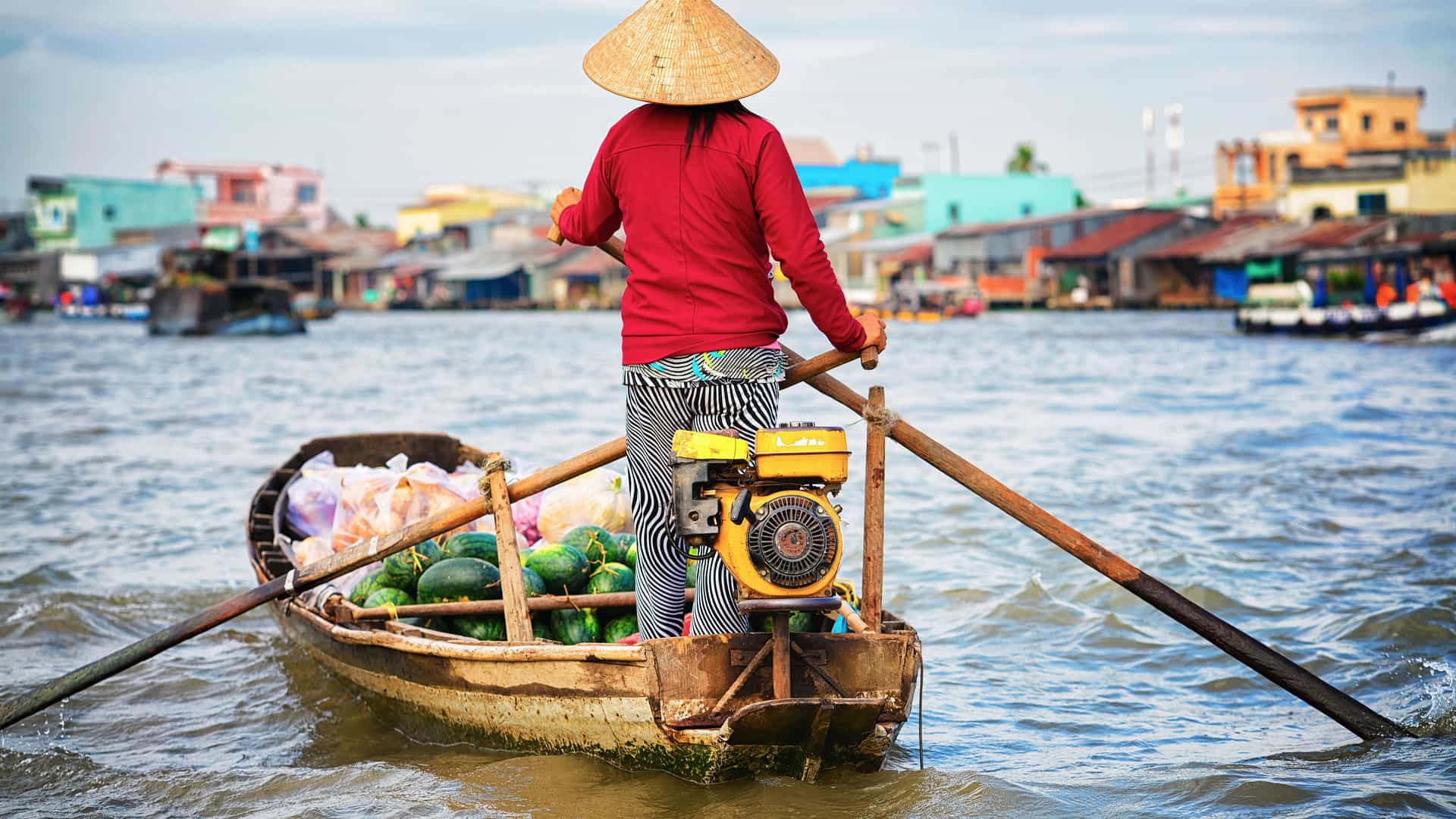 A woman wearing a traditional conical hat paddles a small wooden boat laden with watermelons and other produce on the Mekong River. She is seen from behind, wearing a red long-sleeved shirt and striped pants. The boat is equipped with a small motor. In the background, a bustling floating market and colorful buildings on stilts line the riverbank, capturing a scene from a vibrant floating market in the Mekong Delta, Vietnam.