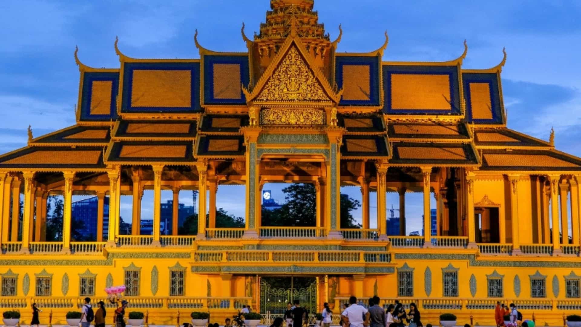 A beautiful view of the Royal Palace in Phnom Penh, Cambodia, at dusk. The golden-yellow building with intricate blue and gold roofs is brightly lit, standing out against the dark blue and orange sky. A small crowd of people is gathered on the grassy area in front of the palace, taking photos and enjoying the evening. The image highlights the detailed and traditional architecture of this Mekong River landmark.