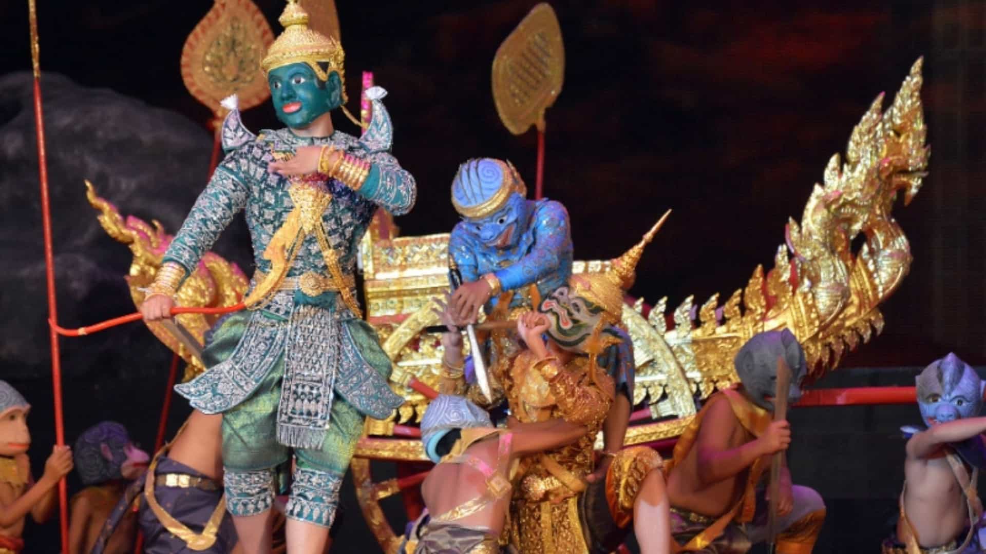 A dynamic performance of a traditional Khmer dance, a form of classical dance from Cambodia. The dancers wear elaborate and colorful costumes and masks, depicting characters from the Ramayana epic. A central figure with green skin and an ornate headdress stands in a dramatic pose. Other dancers in blue and gray masks are positioned around a gilded boat prop. The background is a dark stage, highlighting the vibrant costumes and a rich cultural tradition of the Mekong River region.