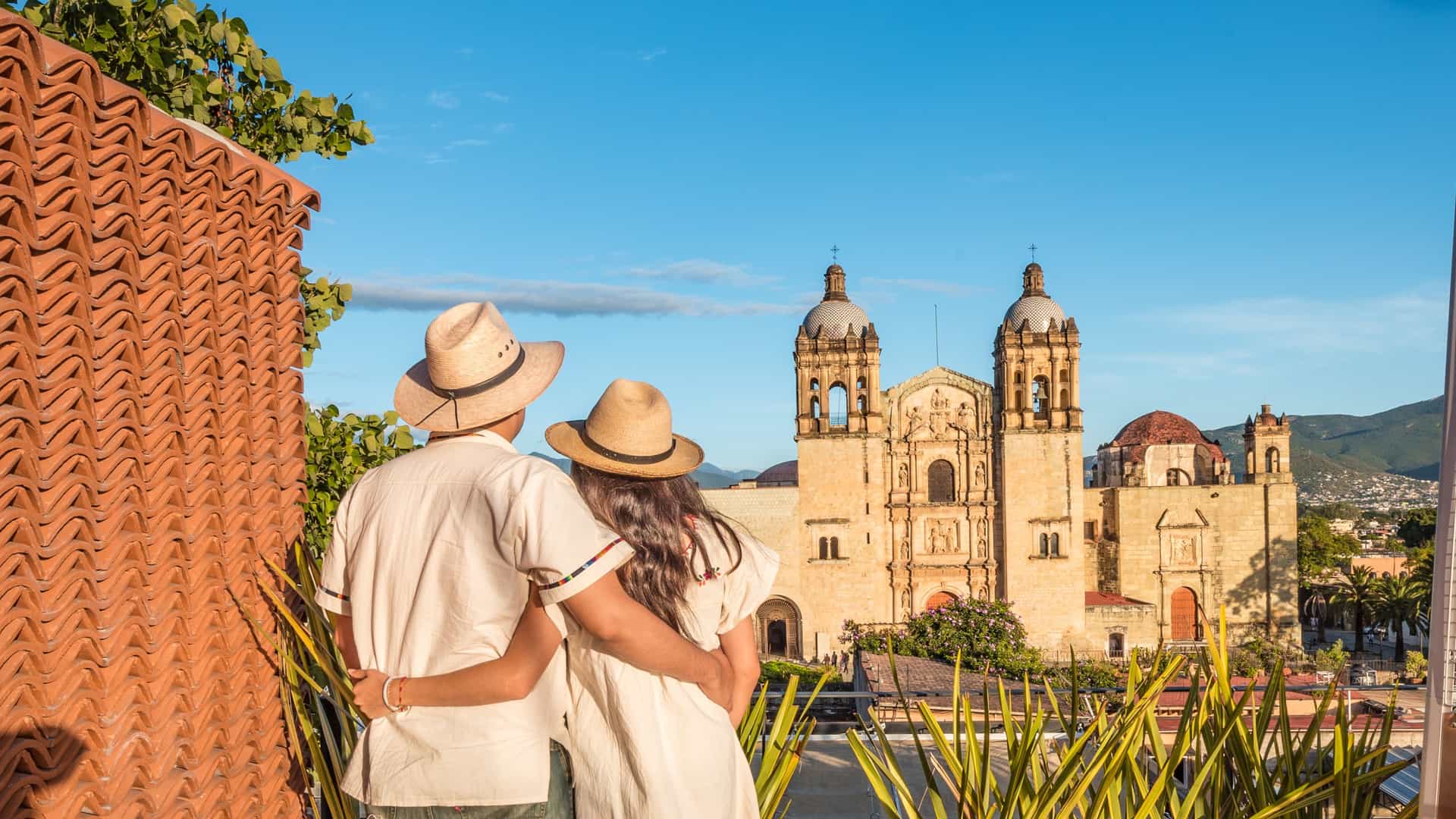A couple wearing straw hats and white shirts stand arm-in-arm on a rooftop, looking out at a grand historic colonial church under a clear blue sky.