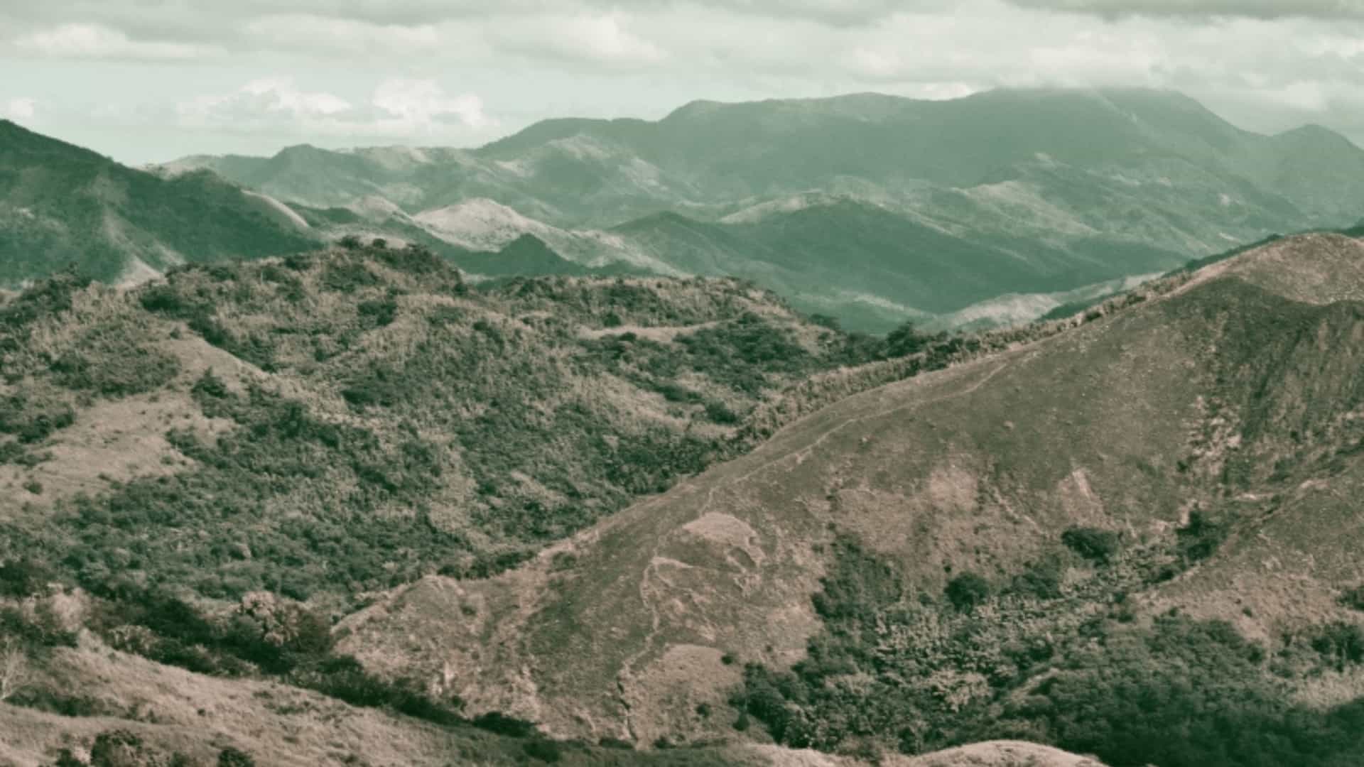An expansive view of the rolling hills and mountains of the Sierra Madre mountain range in Mexico. The landscape is covered in varying shades of green, brown, and gray foliage, with some areas appearing more arid than others. The mountain ridges recede into the distance, creating layers of peaks under a cloudy sky. The image captures the vast and rugged terrain of this prominent Mexican mountain range.