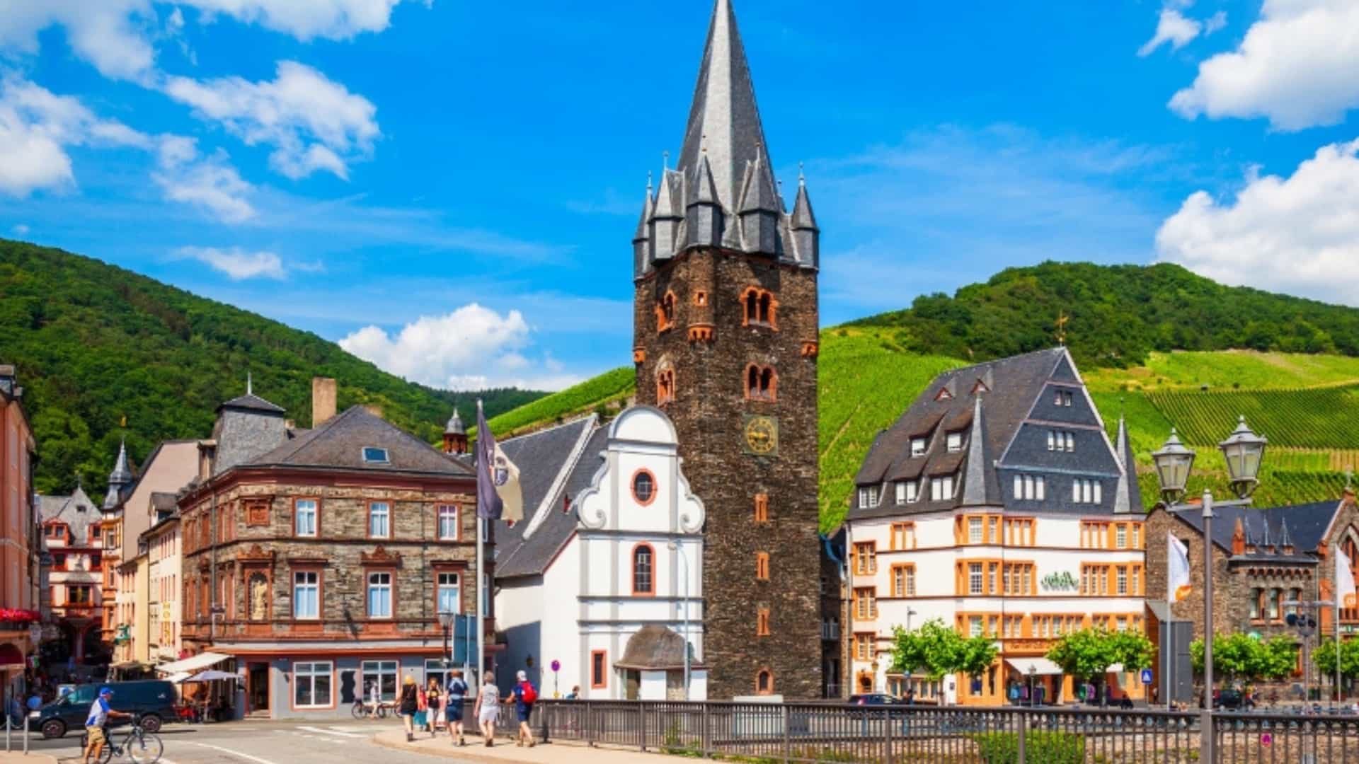 The picturesque town of Bernkastel-Kues, Germany, in the Moselle River Valley. The image shows a street scene with a prominent, tall, Gothic-style stone church tower at its center. Surrounding it are traditional half-timbered houses with colorful facades and slate roofs. In the background, green hills covered with lush vineyards rise under a bright blue sky with white clouds. The scene captures the charm and old-world character of this famous wine region.