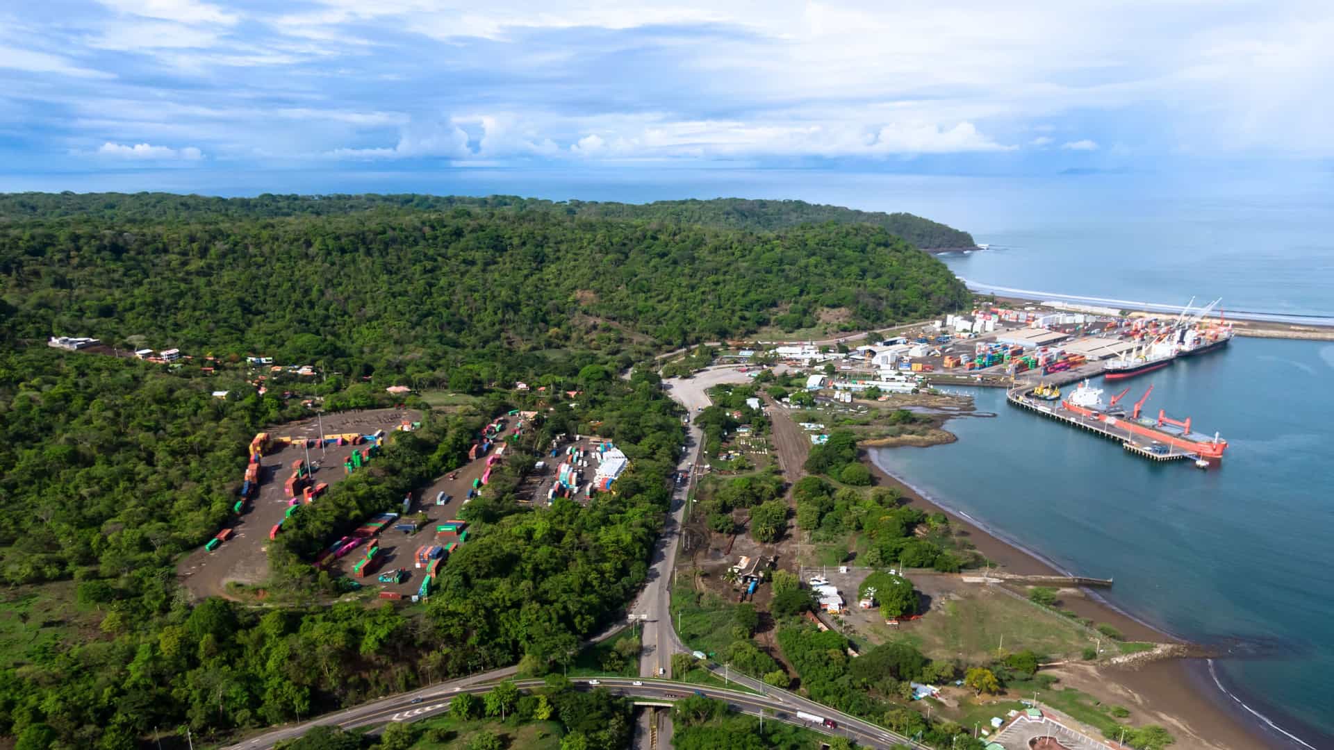 Aerial view of Caldera Port in Puntarenas, Costa Rica, showing cargo docks, shipping containers, and surrounding forested coastline.