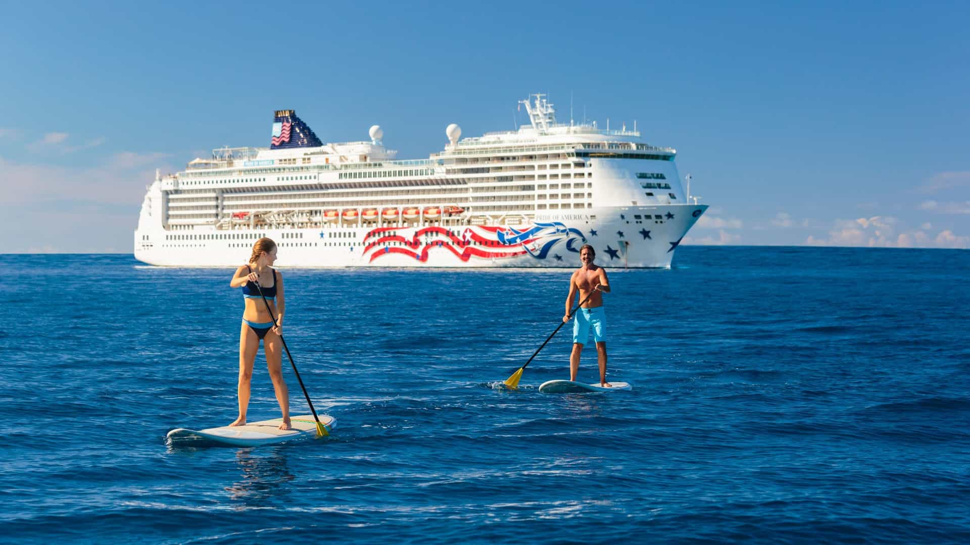 A man and a woman are stand-up paddleboarding on a body of water with the Norwegian Cruise Line's Pride of America ship in the background on a sunny day.
