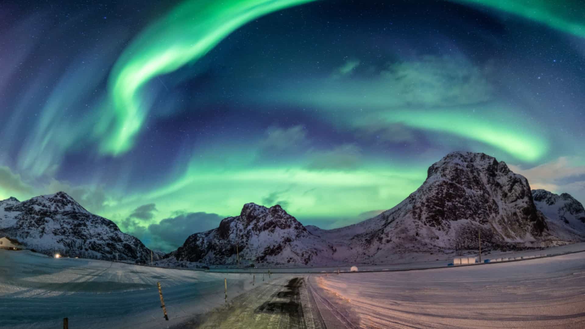 A spectacular display of the green Aurora Borealis arching over snow-capped mountains and a road within the Arctic Circle in Norway, a view often seen on an Oceania cruise.