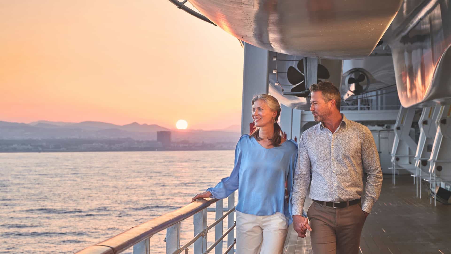 A couple enjoys a romantic sunset stroll on the deck of an Oceania Cruises ship, gazing at the horizon during a serene Trans-Atlantic crossing.