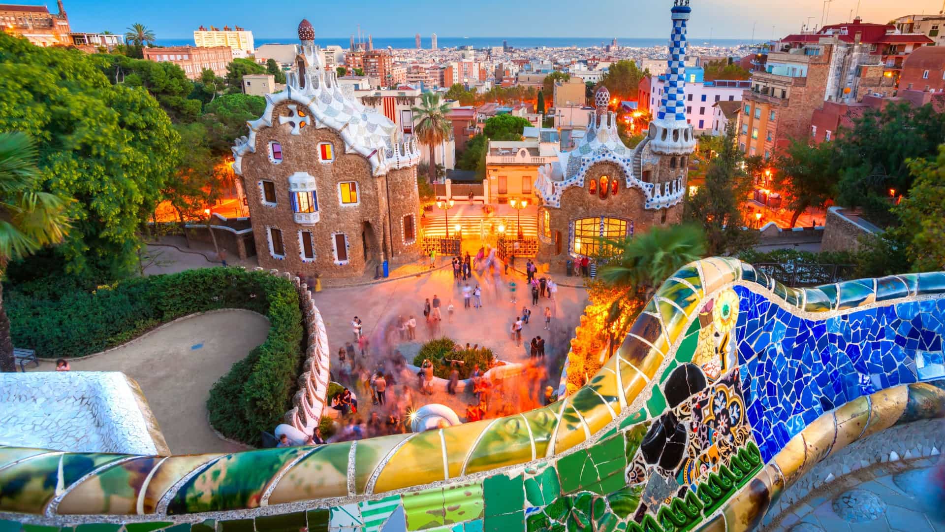 Sunset view of Antoni Gaudí's Park Güell in Barcelona featuring distinctive white and blue mosaic-tiled gatehouses with ornate details, the main plaza with visitors, lush greenery, palm trees, and the city's colorful buildings and Mediterranean coastline stretching across the horizon.
