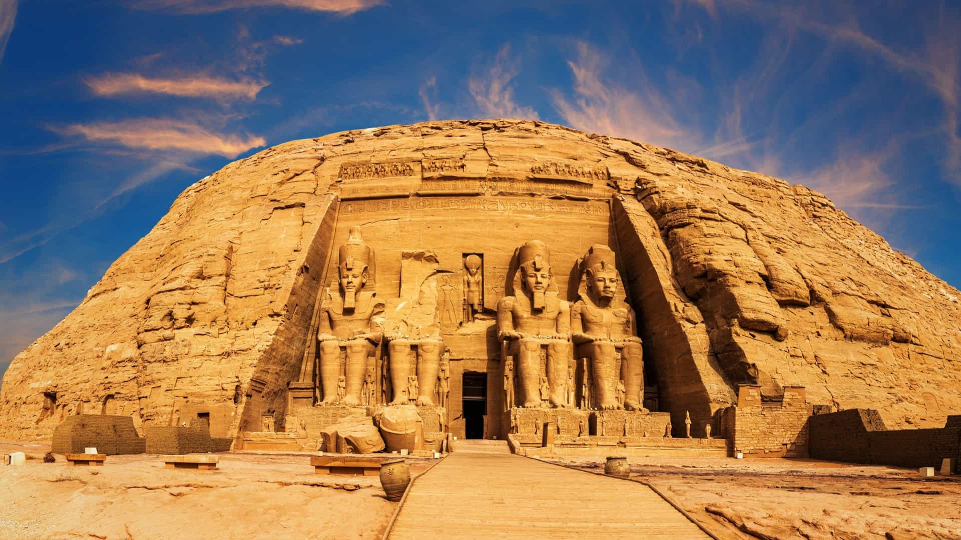 A magnificent exterior view of the Great Temple of Ramesses II at Abu Simbel, Egypt, featuring four colossal seated statues carved into the rock under a dramatic sky.