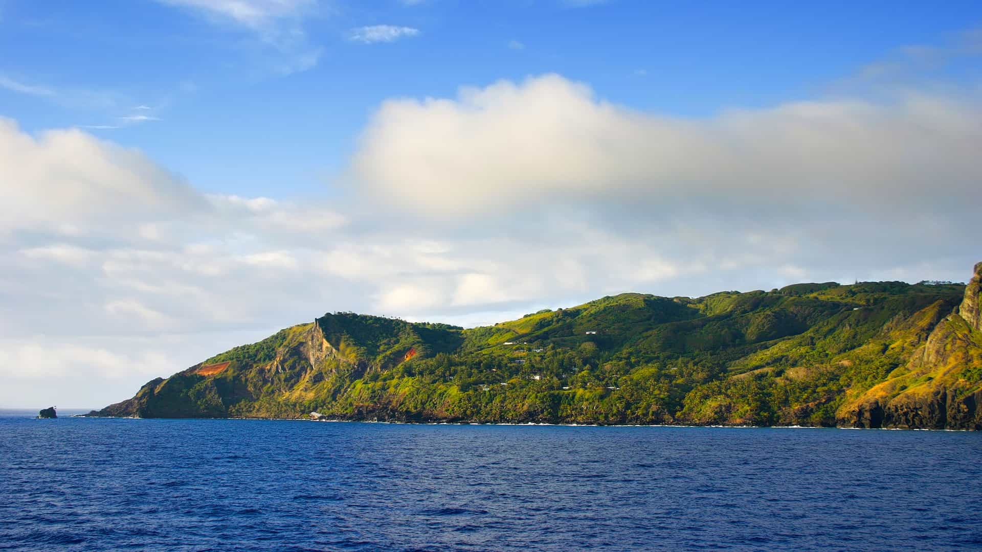 A panoramic view of the rugged coastline of Pitcairn Island, with lush green hills and cliffs meeting the deep blue Pacific Ocean.