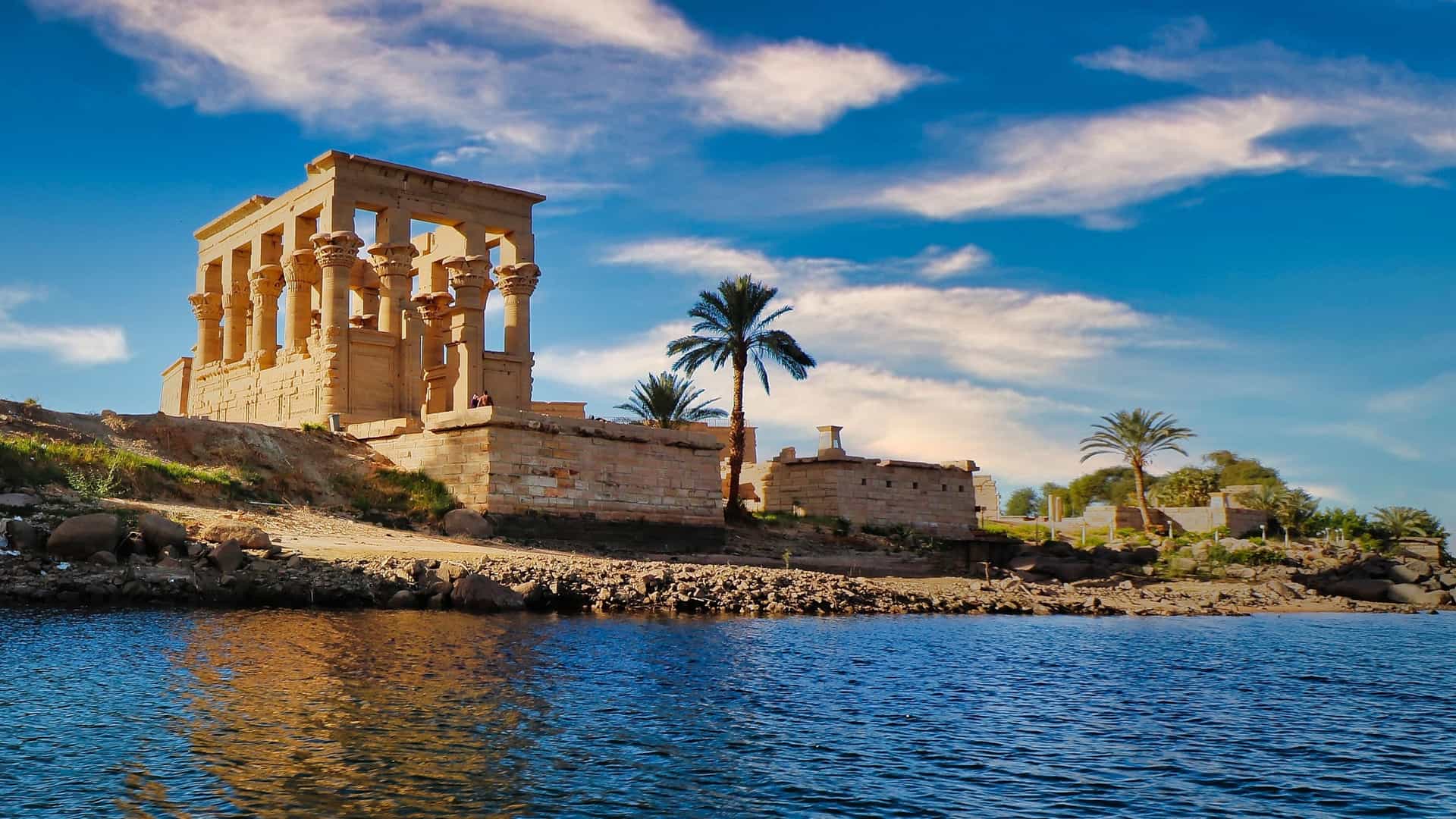 A wide-angle view of the ancient Temple of Philae on Agilkia Island, with its intricate columns and a palm tree reflected in the waters of the Nile River.