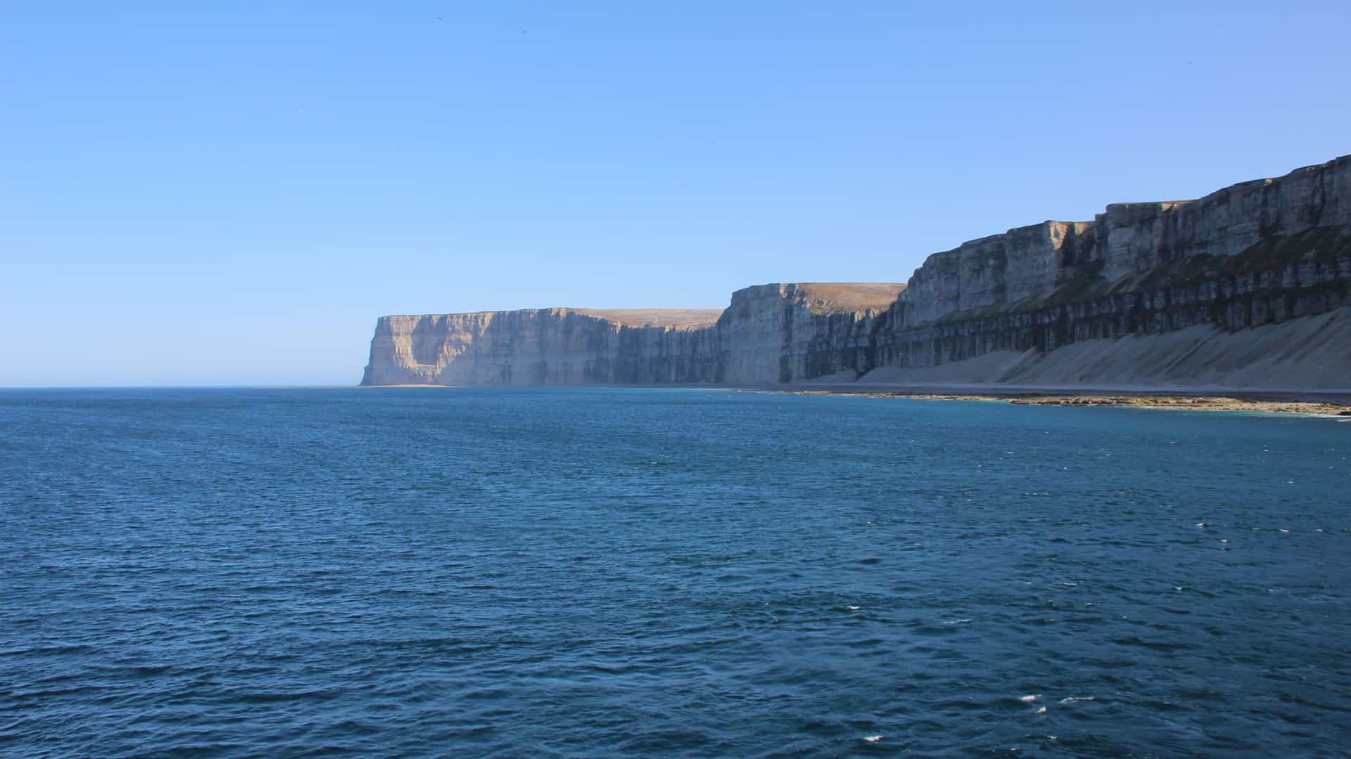 A dramatic view of the sheer limestone cliffs of Akpatok Island, towering over the deep blue sea under a clear, bright sky.