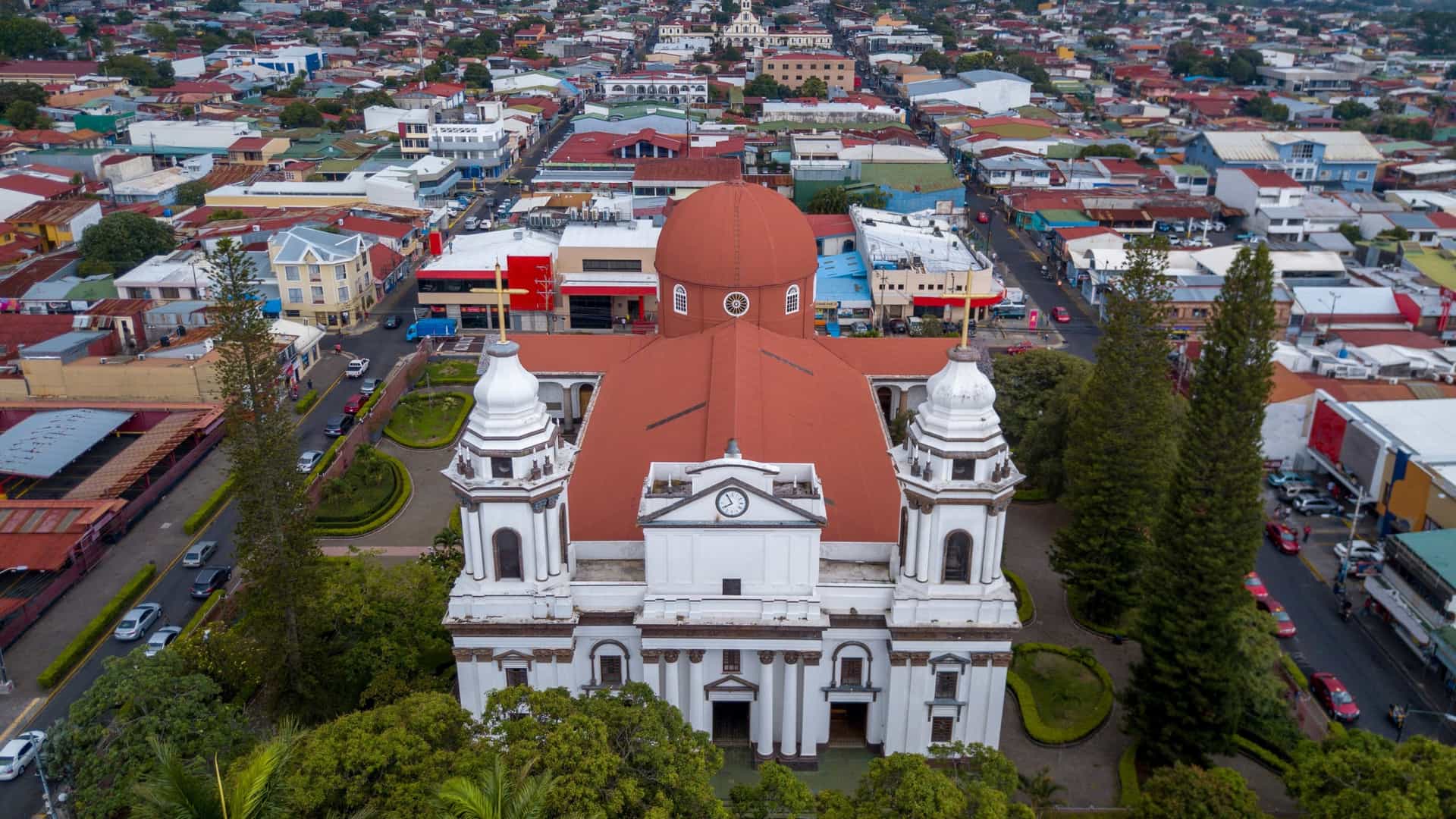 An aerial view of the city of Alajuela, Costa Rica, dominated by the central white and red-domed structure of the Catedral de Alajuela (Alajuela Cathedral), surrounded by the urban landscape.