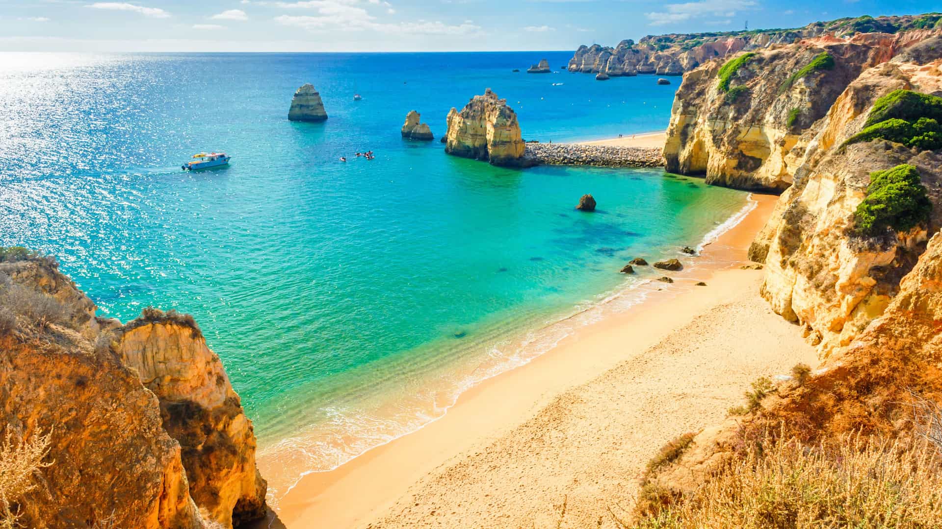 A stunning view of a small, golden-sand beach on the Algarve Coast, Portugal, nestled between towering, eroded sea cliffs. The calm, clear water of the bay is a vibrant turquoise, with several dramatic rock stacks rising from the ocean. A small boat is anchored in the water under a clear, bright blue sky.