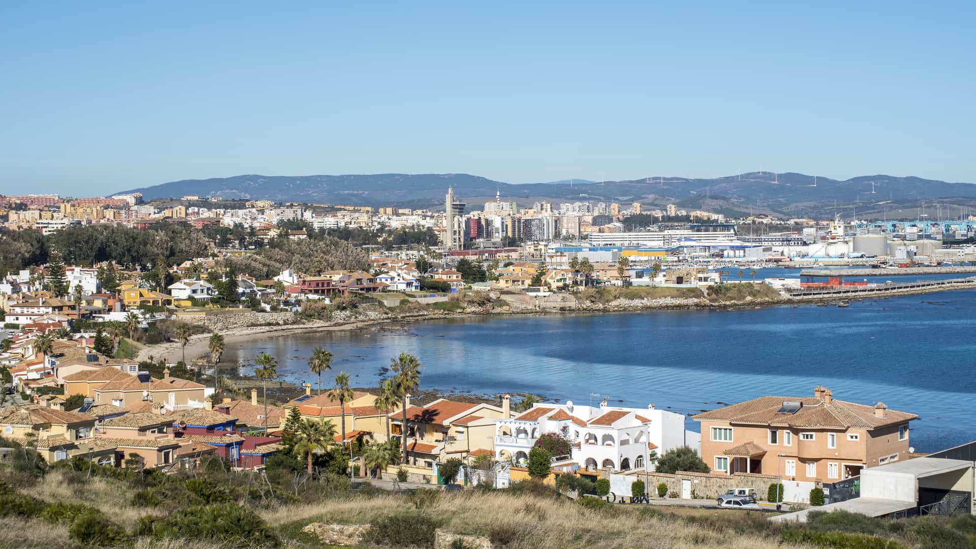 An elevated, panoramic view of the coastal city of Algeciras, Spain, and its bay. The calm blue water is bordered by a shoreline with residential houses, while the main city and a large industrial port with cranes and ships are visible in the background. Hills with wind turbines can be seen in the distance under a clear blue sky.