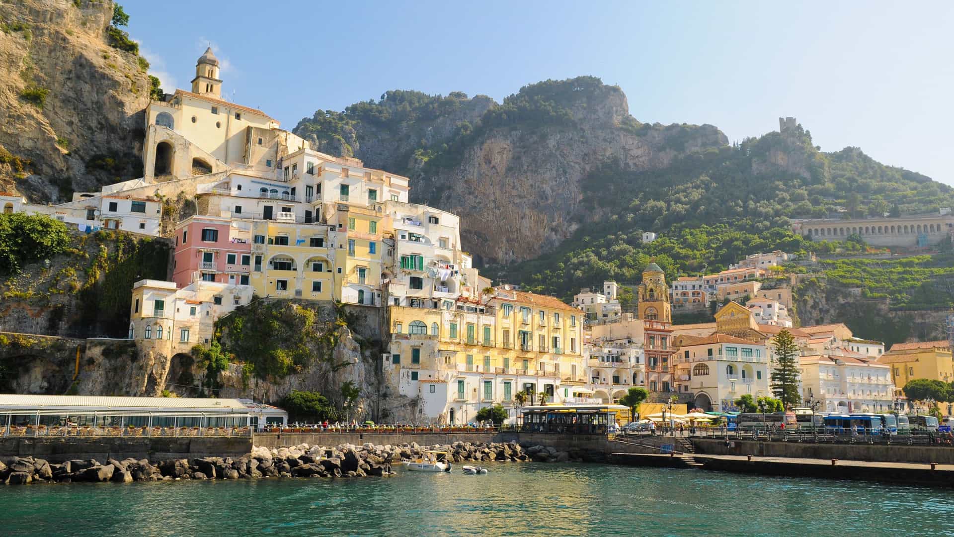 A sunny waterfront view of the colorful, cliffside village of Amalfi in Italy, with historic buildings and mountains rising from the Tyrrhenian Sea.
