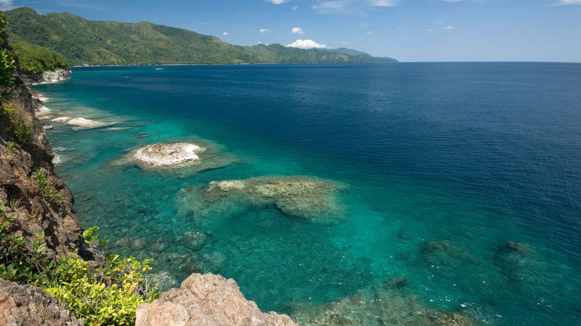 A breathtaking tropical landscape of Ambon Island, Indonesia, with a view of a lush green coastline and the clear turquoise waters of the Banda Sea revealing a shallow coral reef.