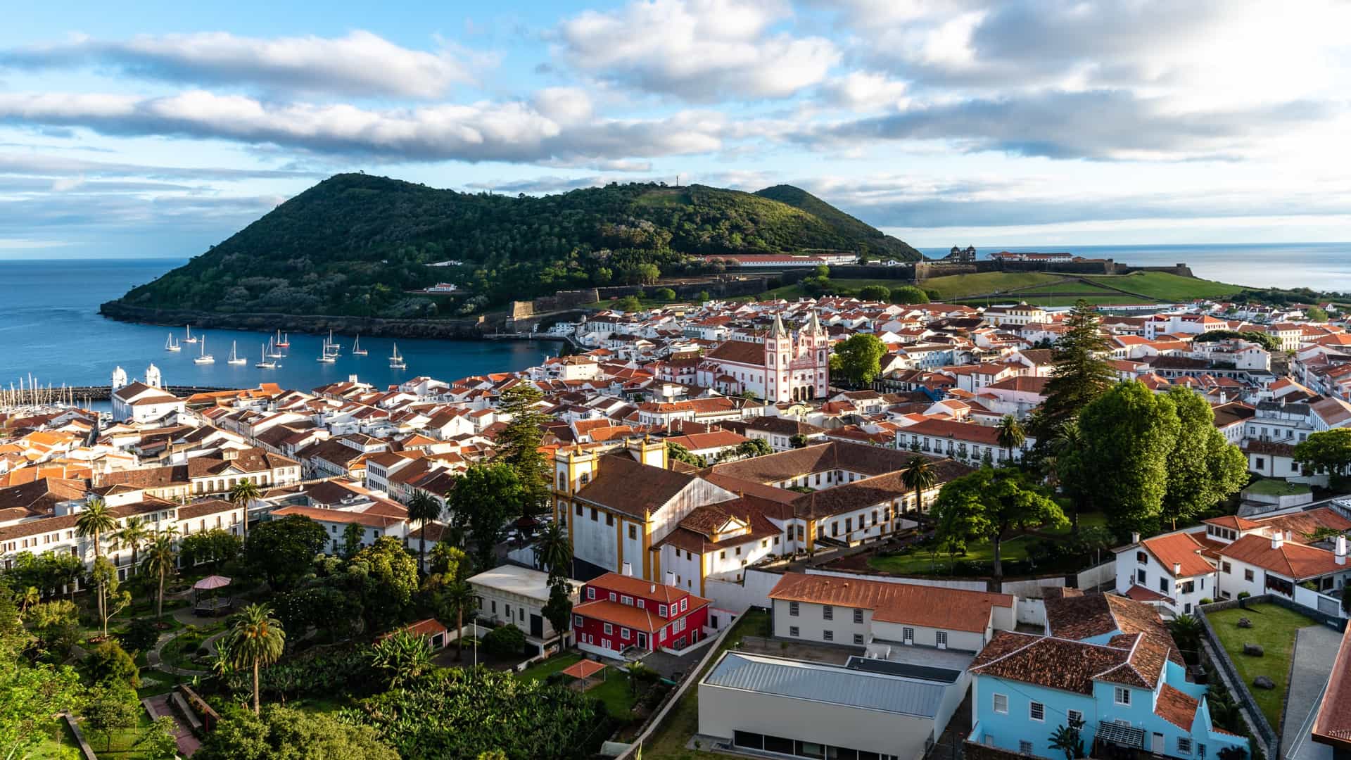 A coastal town in Angra do Heroísmo with white buildings and red roofs, a historic yellow‑and‑white landmark in the foreground, a small park, and a calm bay with sailboats backed by a forested hill and fortress.