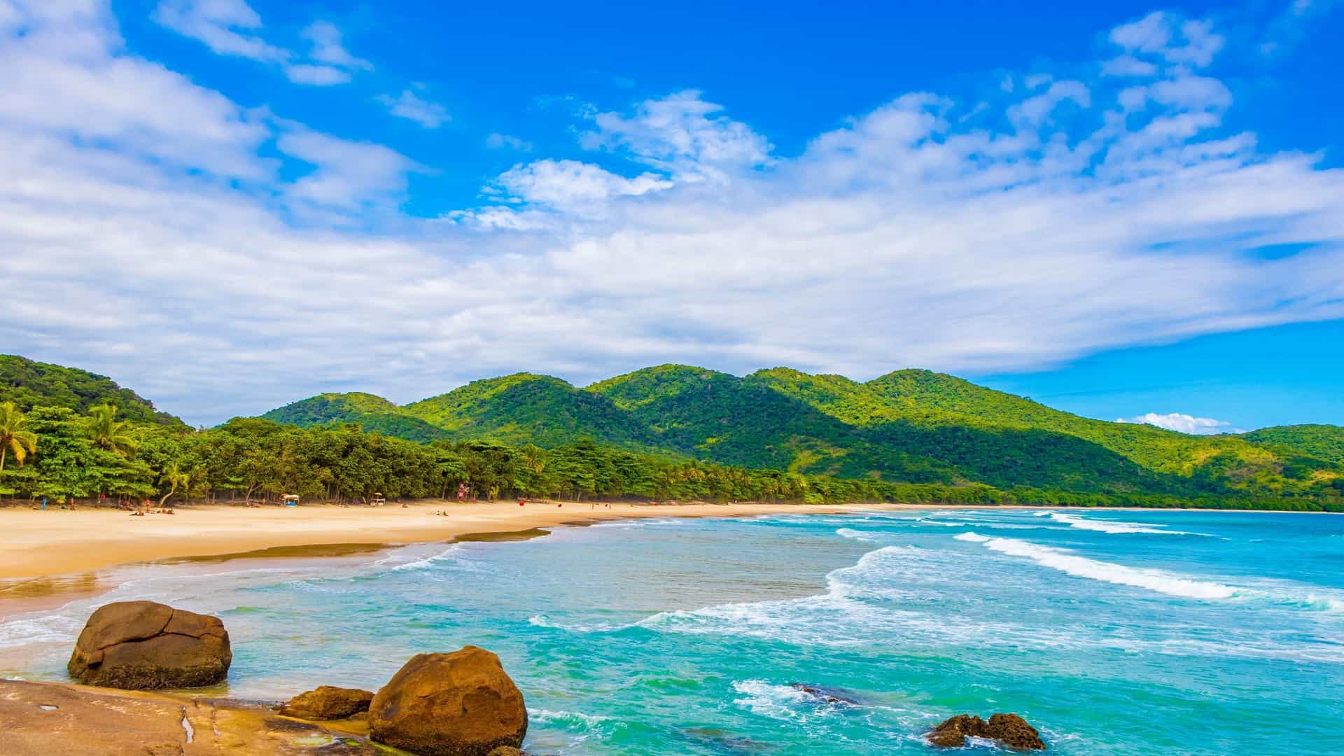 An aerial or wide view of Praia de Lopes Mendes, a pristine, crescent-shaped beach on Ilha Grande, Brazil. The shoreline features a long stretch of fine, powdery white sand meeting bright, clear aquamarine water. The entire beach is backed by the dense, lush Atlantic rainforest, with gentle waves from the open ocean rolling onto the shore.