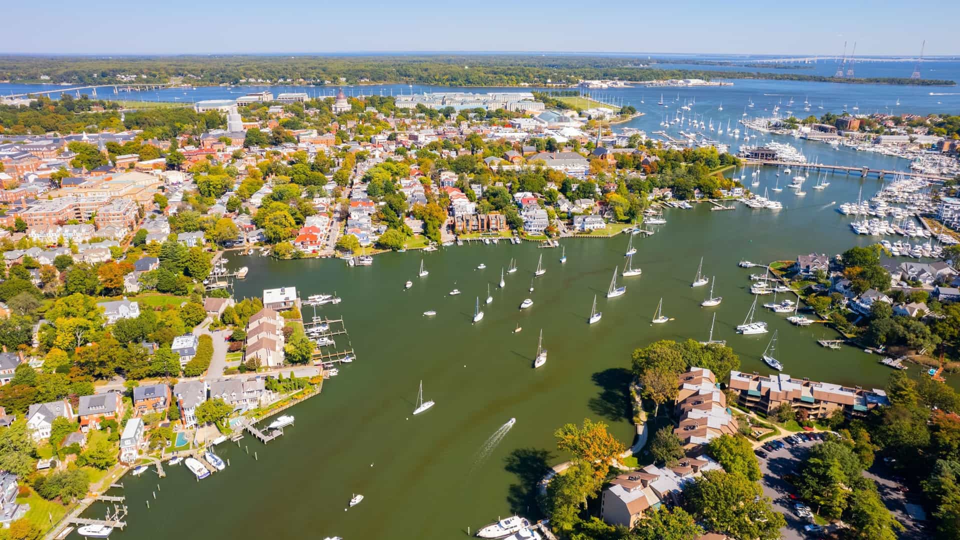 An aerial, high-angle view of a busy Maryland harbor, likely in Annapolis. The image shows numerous docked pleasure boats and ships floating in the water, with waterfront properties, docks, and piers visible along the curved shoreline. The harbor is bordered by buildings and streets of the coastal city under daylight.