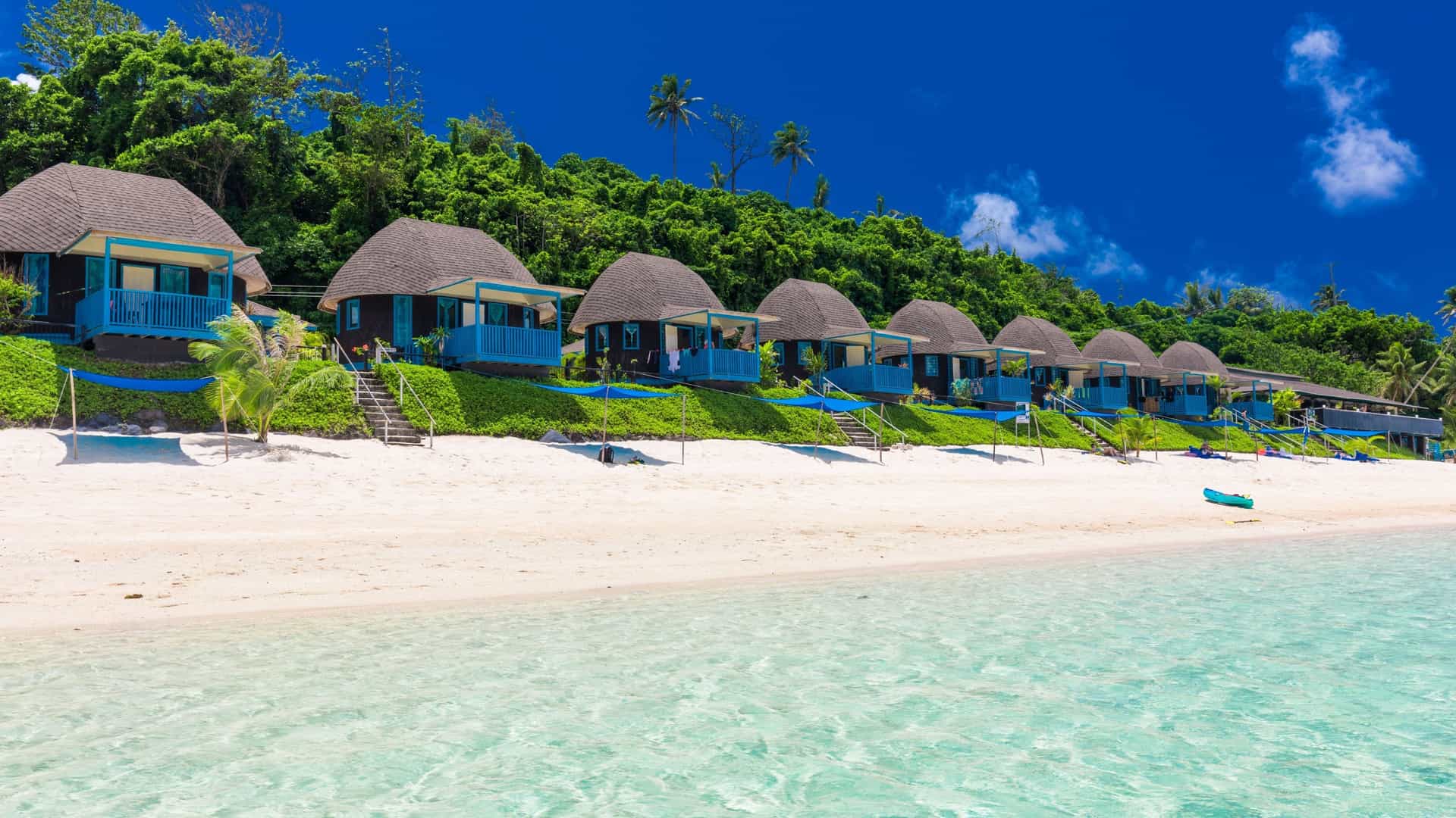 A scenic shot of a line of small, blue, thatched-roof beach bungalows on a pristine white sand beach in Apia, Samoa, with a clear turquoise ocean and a lush green hill in the background.