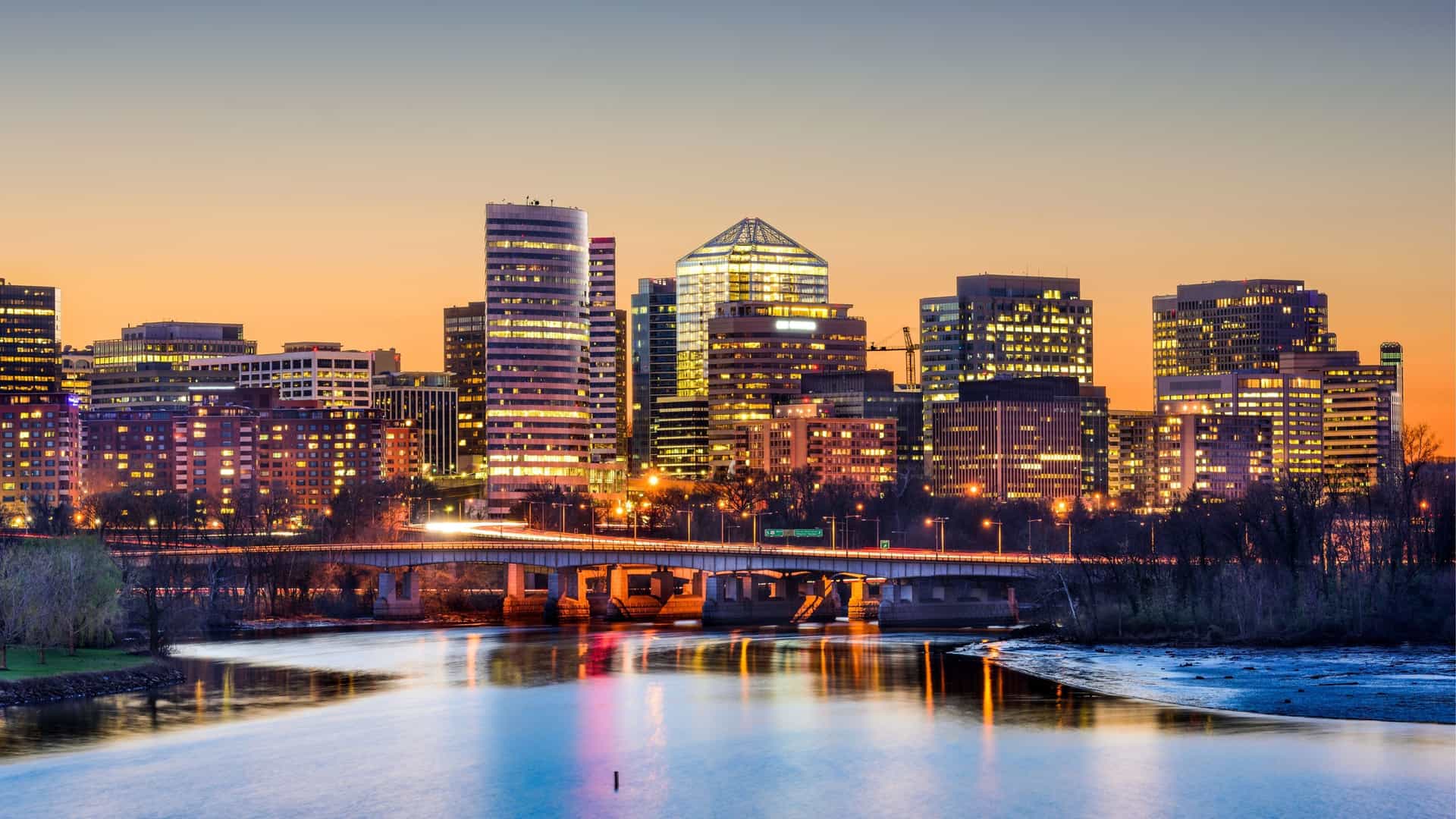 The illuminated skyline of Arlington, Virginia, glows at sunset, reflected in the river below a bridge.