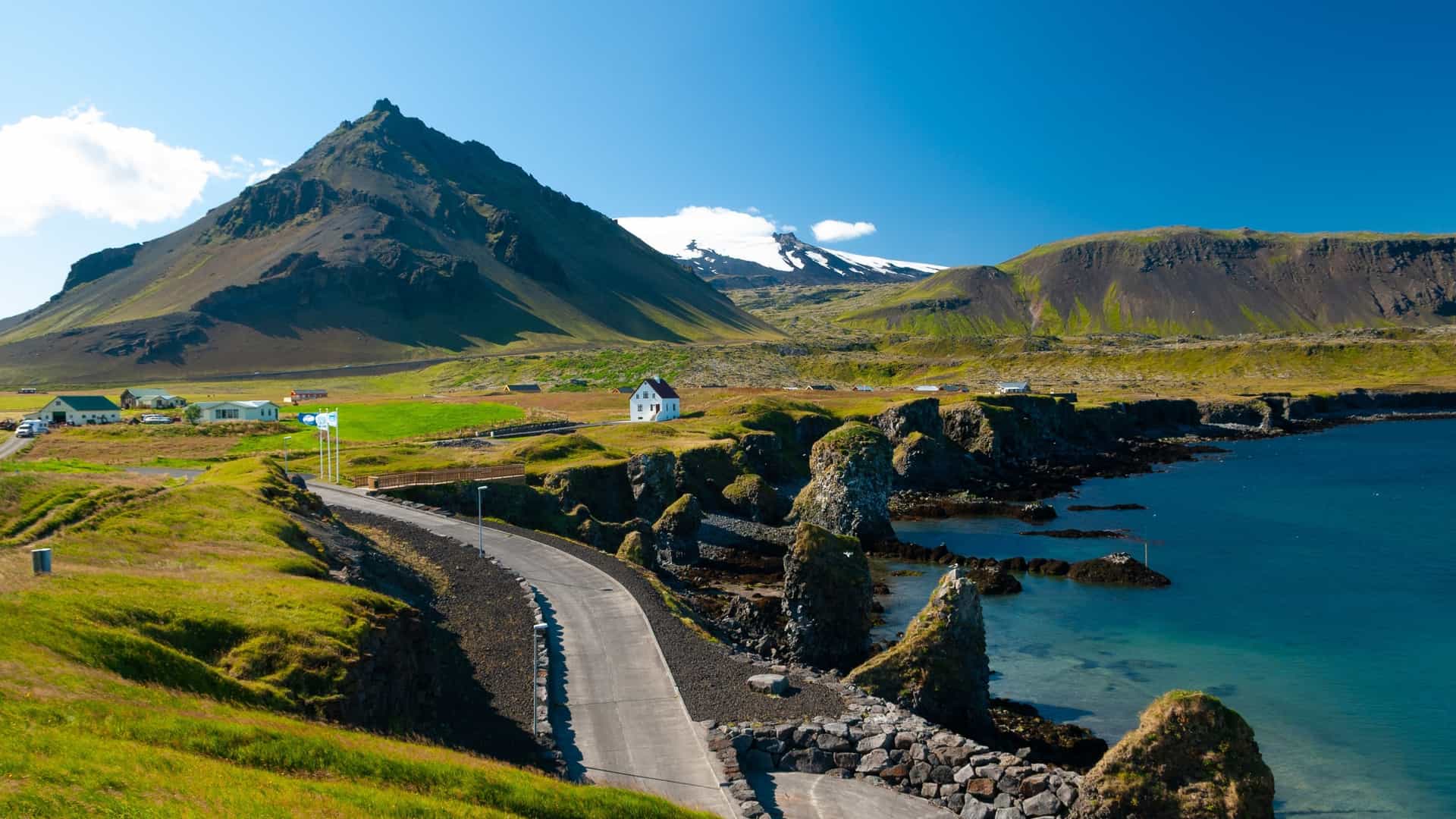 A scenic road curves along the coast near Arnarstapi, Iceland, with dramatic cliffs, a white house, and a snow-capped mountain backdrop.
