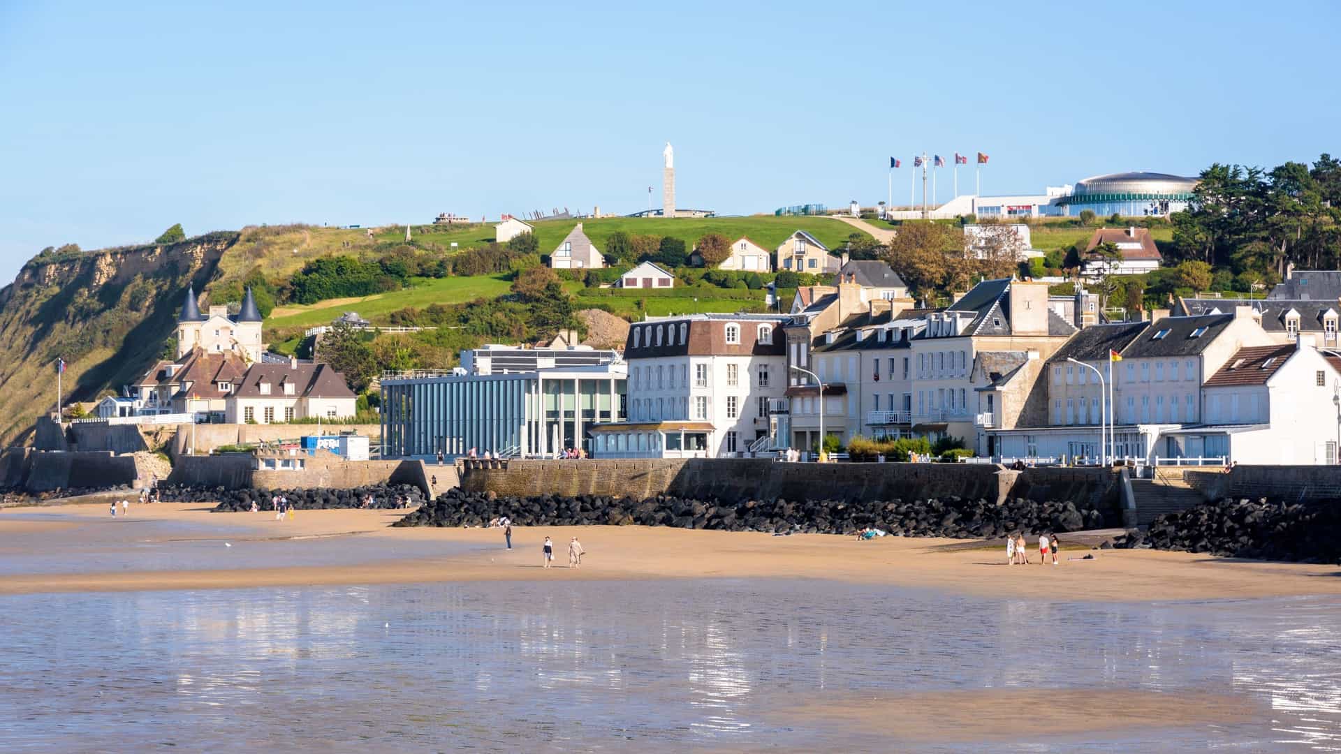 A scenic view of Arromanches-les-Bains in Normandy, France, featuring the sandy beach, historic buildings along the waterfront, and a hilltop museum under a clear blue sky.