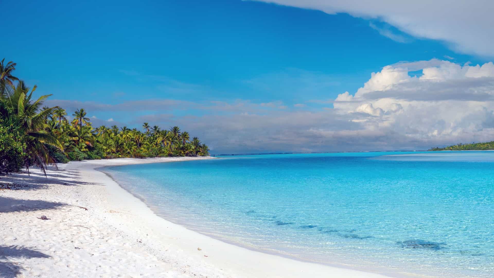 A serene white sand beach with palm trees in Arutanga, Cook Islands, with crystal-clear turquoise water stretching out to the horizon under a blue sky with fluffy white clouds.