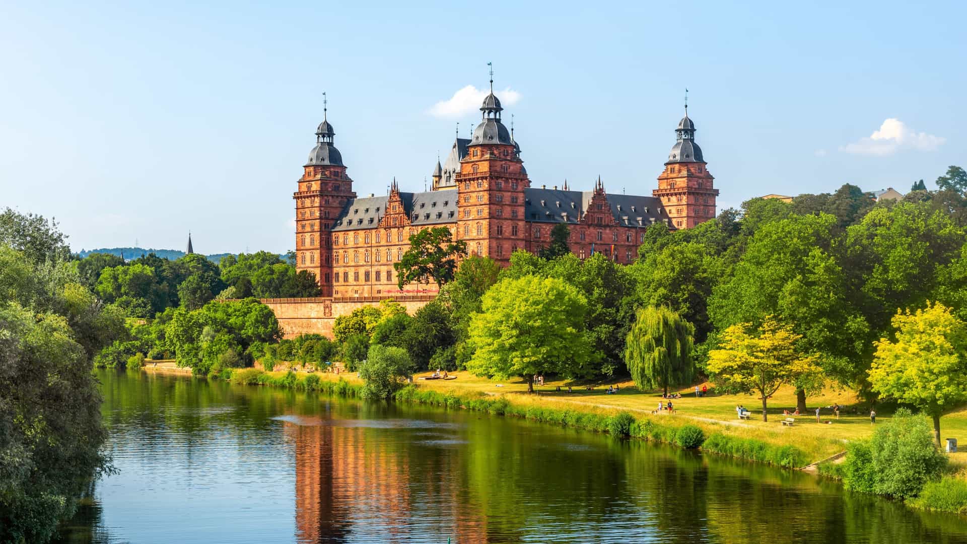 The red sandstone Johannisburg Palace stands majestically along the banks of the Main River surrounded by lush green trees in Aschaffenburg, Germany.