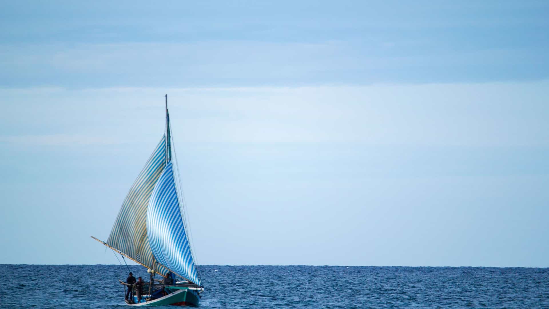 A sailboat with blue and white striped sails sailing on the calm blue waters of the Indian Ocean near Ashmore Reef, Australia, on a hazy day.