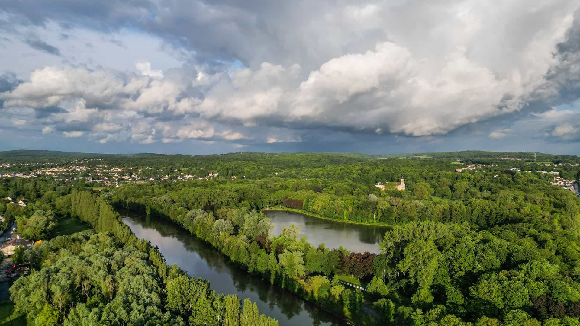 An aerial view of the Oise River cutting through lush green woodlands in Auvers-sur-Oise, France, under a dramatic, cloudy sky.