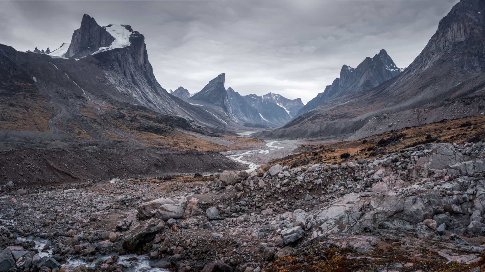 A stunning panoramic view of the rugged, mountainous landscape of Auyuittuq National Park in Nunavut, Canada, featuring a river winding through a rocky valley under a dramatic sky.